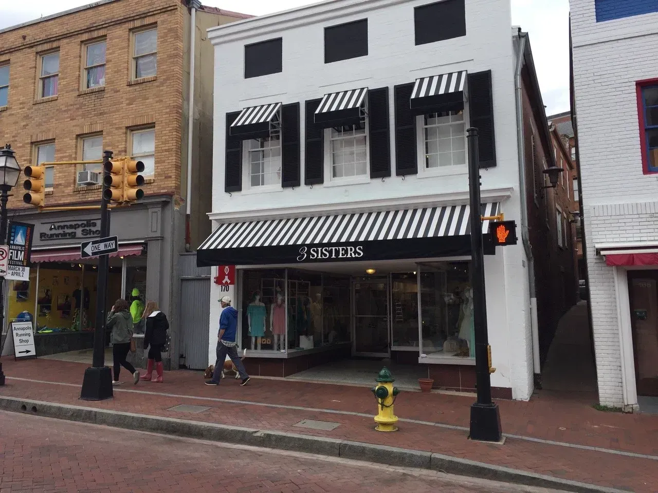 Street view of a white storefront with black awnings, a sidewalk, pedestrians, and a traffic light.