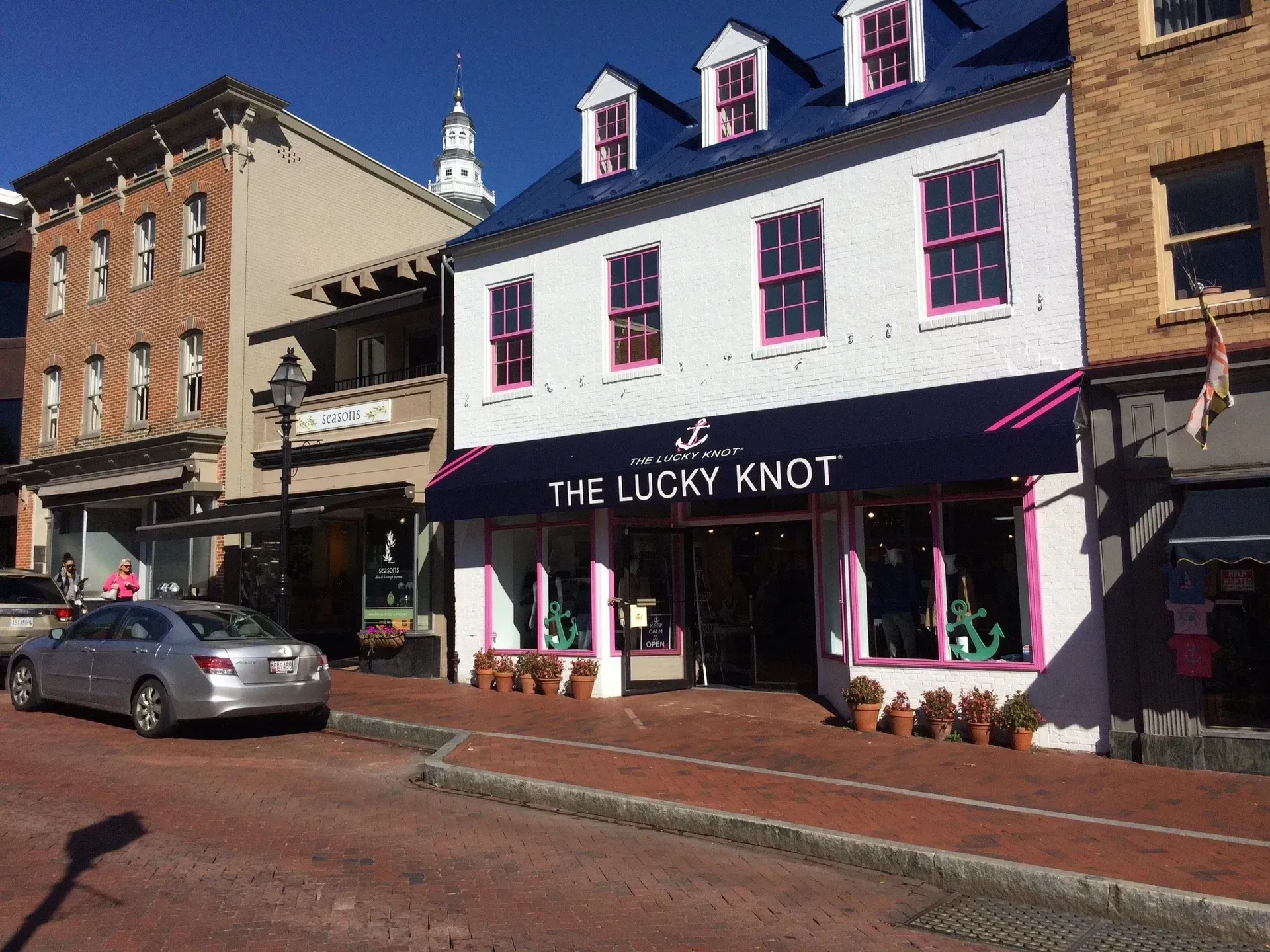 Street view of The Lucky Knot shop with white and pink facade on a sunny brick-lined main street