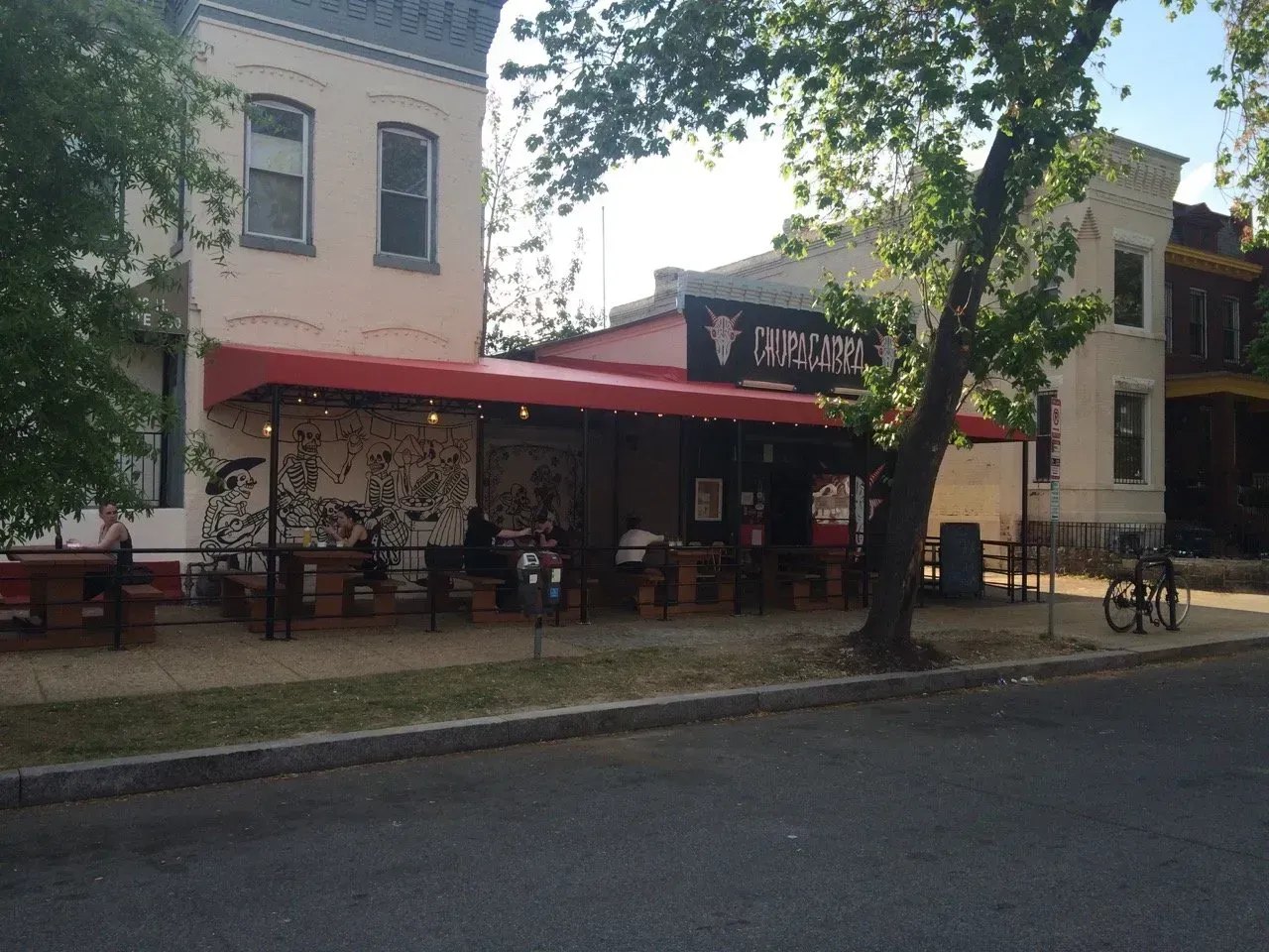 Small town storefront with red awning, trees, and outdoor seating along a quiet street.