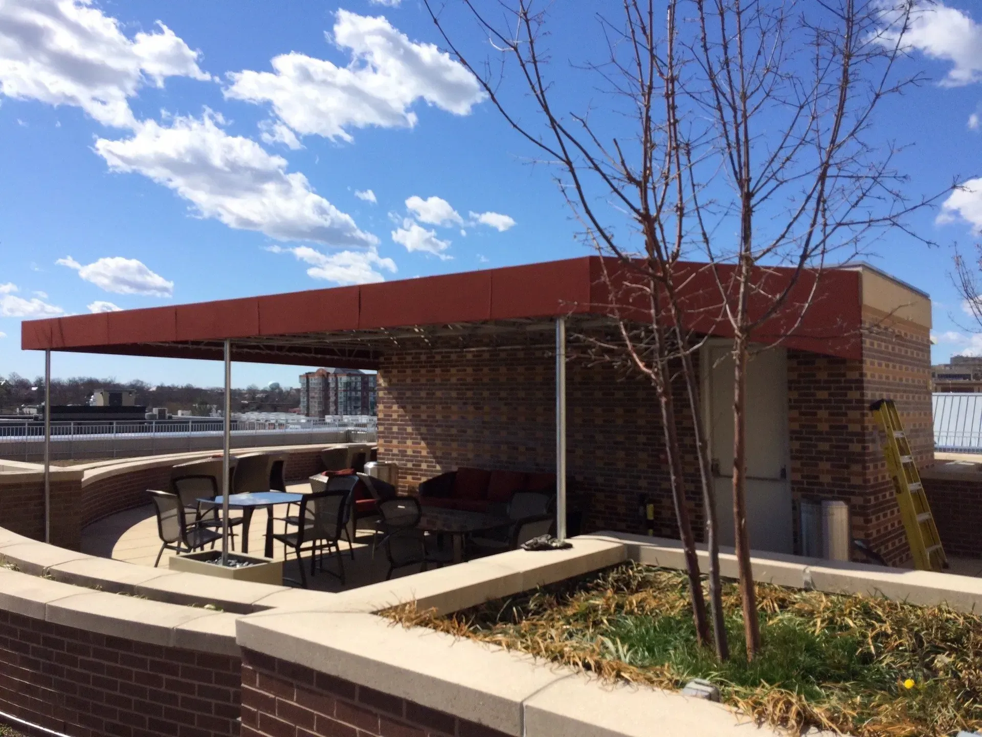Outdoor café patio with tables under a red canopy beside a curved concrete wall on a sunny day