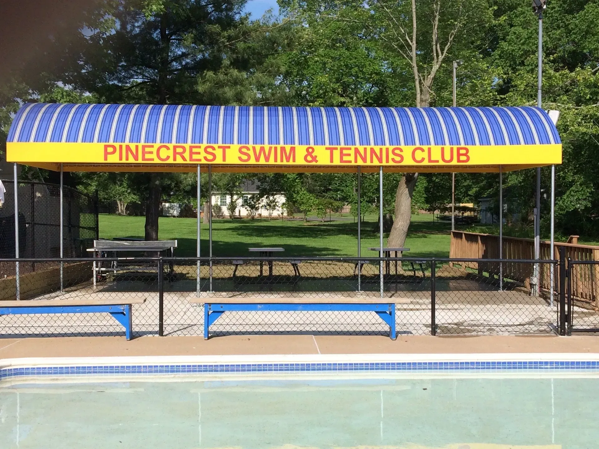 Pinecrest Swim & Tennis Club sign under a blue-and-yellow awning beside a pool and fence