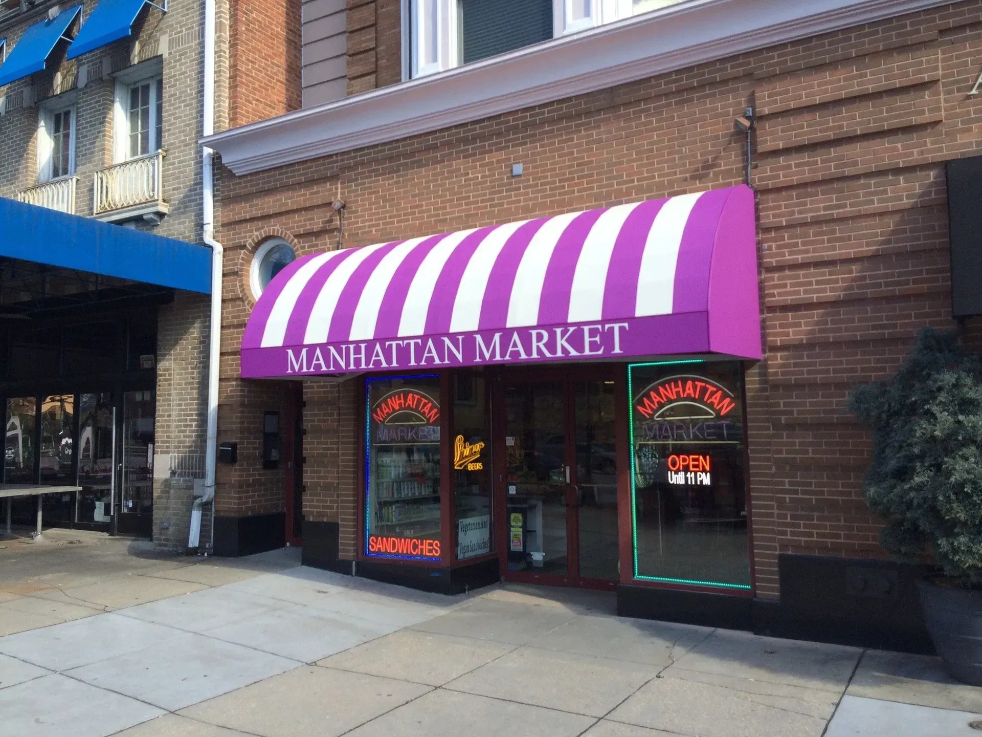 Manhattan Market storefront with purple striped awning and red neon open sign on a city sidewalk
