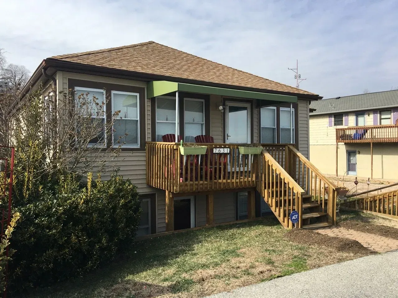Small house with a front deck, stairs, and railing in a grassy yard under a tiled roof.
