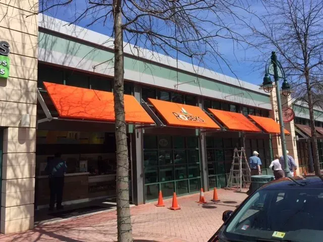 Storefront with orange awnings and glass entrance, cones on sidewalk, and a few people standing outside.