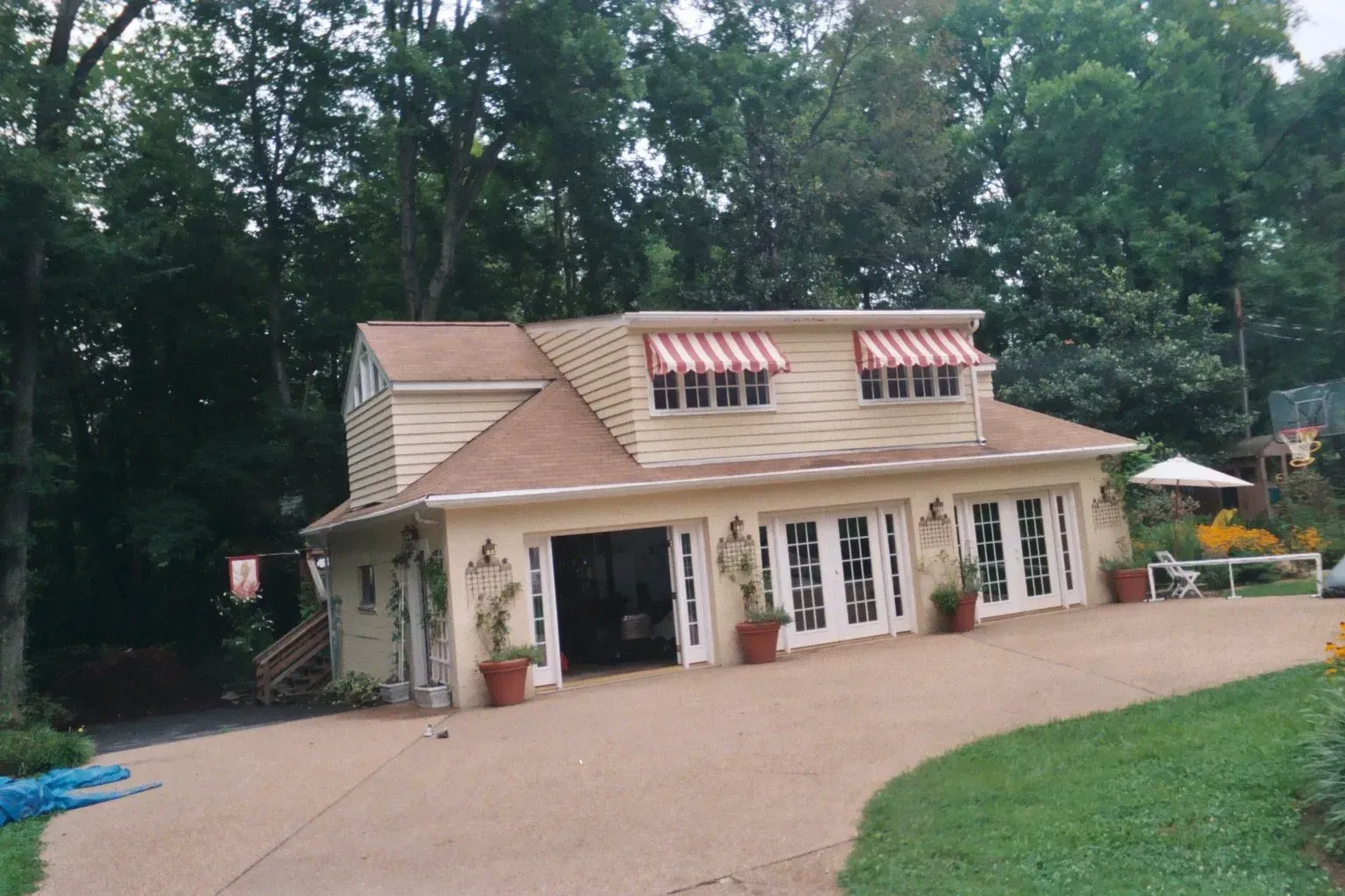 Cream-colored cottage with tan roof and dormer windows, set beside a gravel drive and trees.