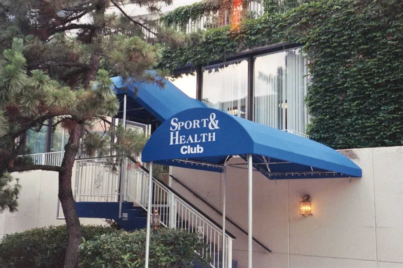 Blue awning for Sport & Health outside a building amid trees and stairs