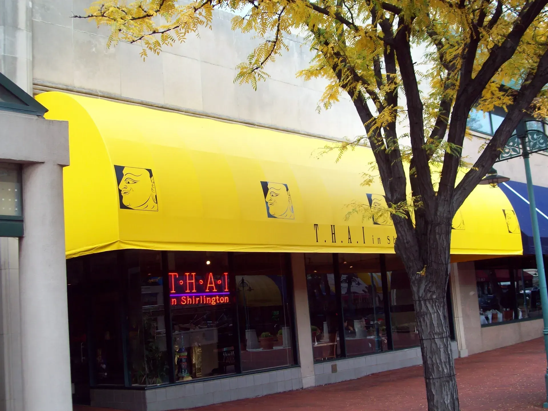 Storefront with bright yellow awning, tree in front, and neon open sign in the window.