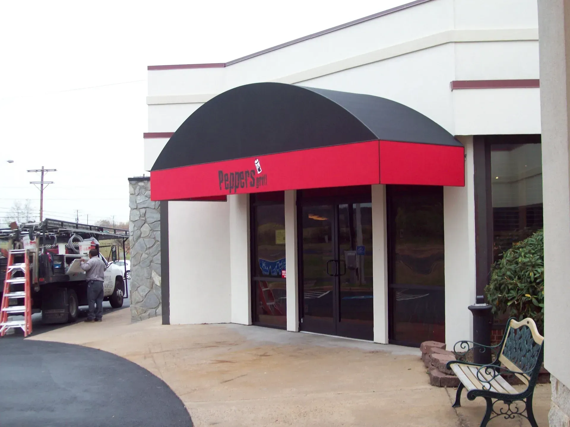 Storefront entrance with red awning and glass doors beside a bench and parked vehicles