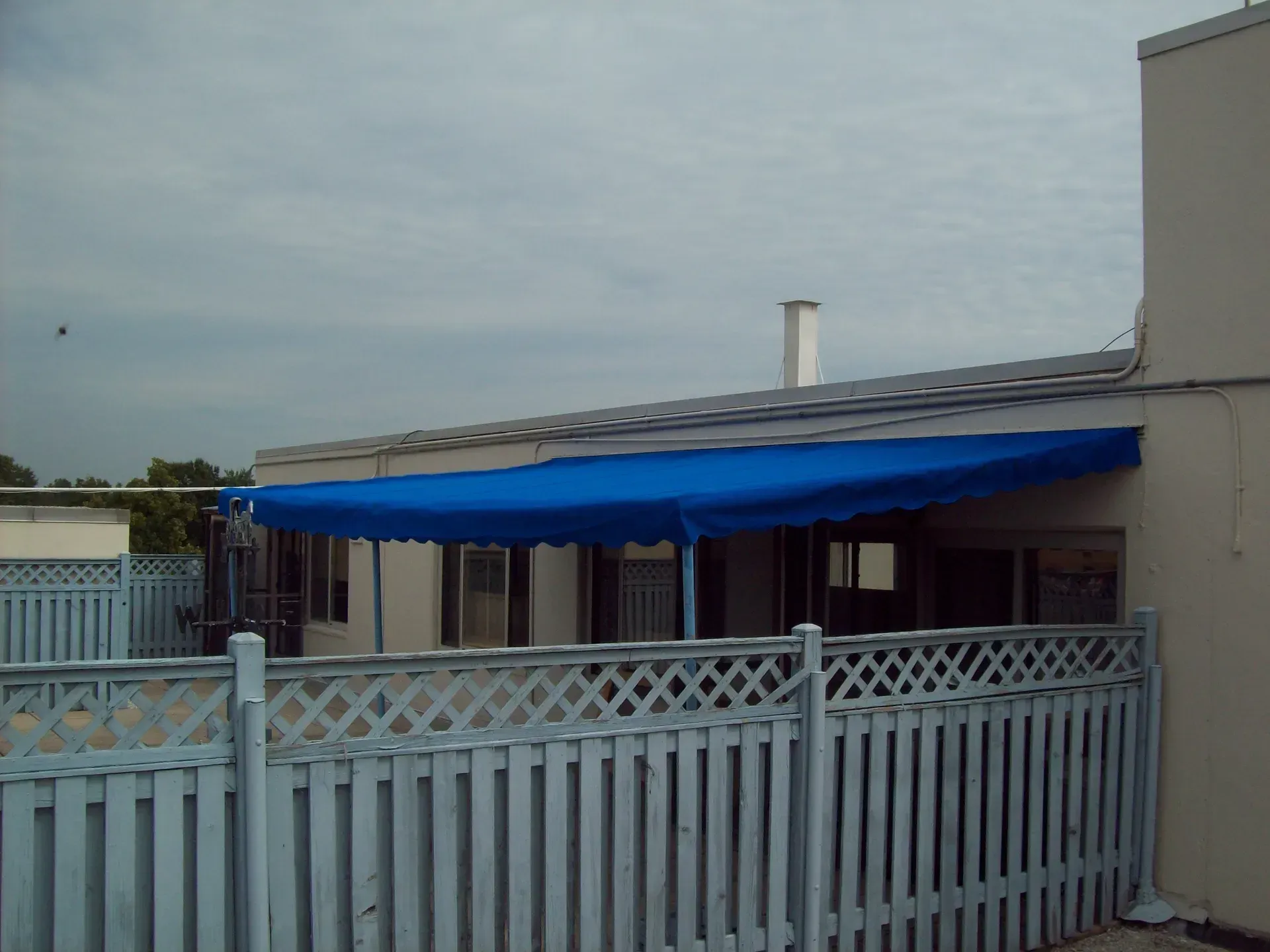 Backyard patio with a blue awning behind a white fence under a gray sky