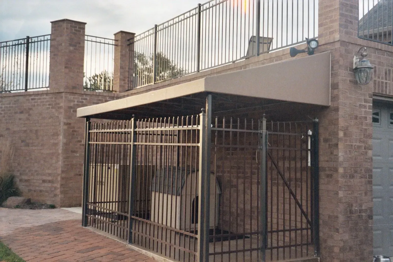 Outdoor stairwell enclosed by a metal cage beneath a brick building with a fenced rooftop.