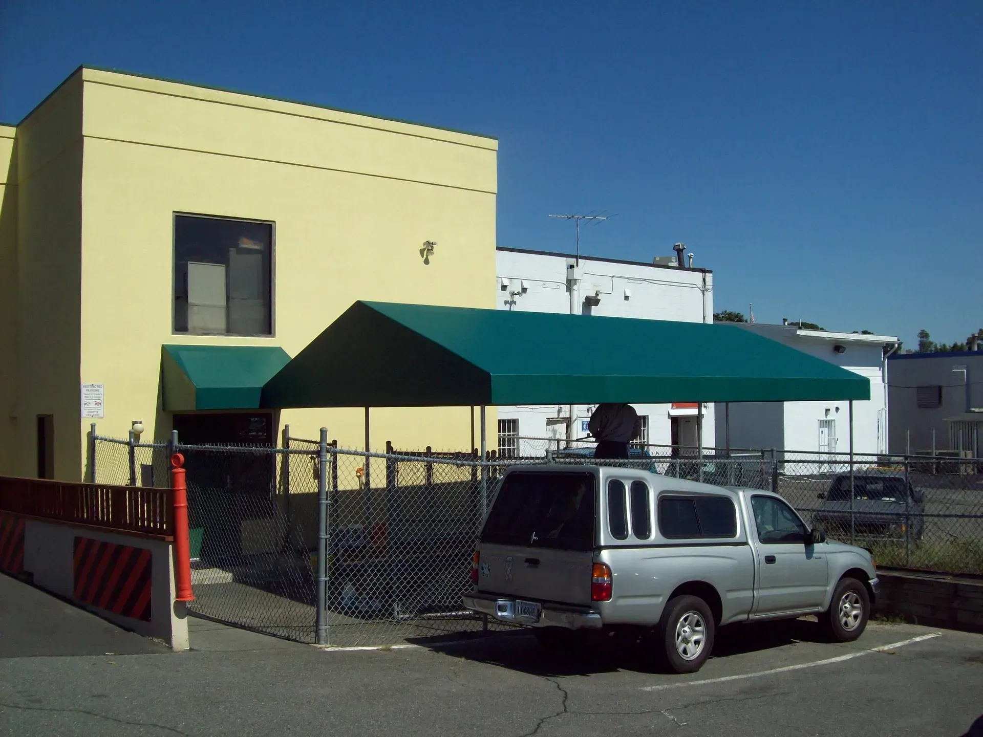 Yellow building with green awning, chain-link fence, and parked silver pickup truck under a blue sky.