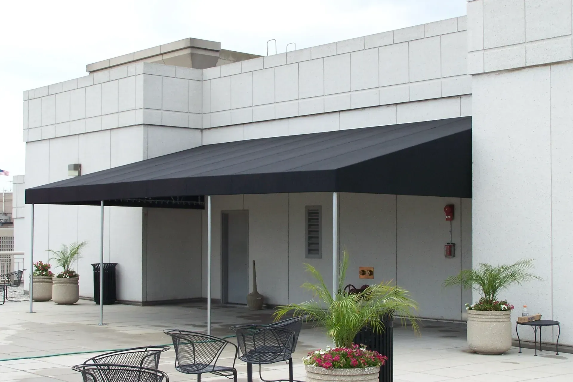 Modern white building with a black awning, patio tables, potted plants, and a flower planter in front.