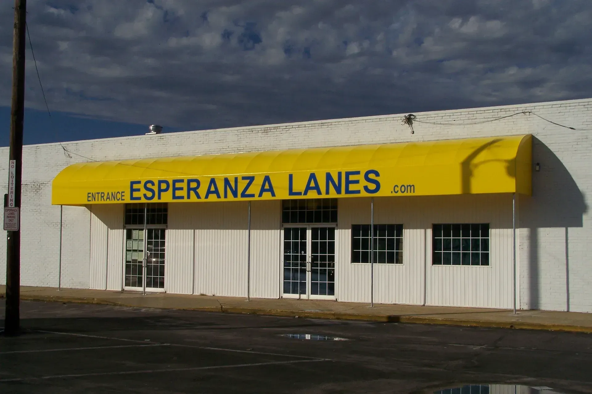 White storefront with yellow awning reading “Esperanza Lanes” under a dark cloudy sky