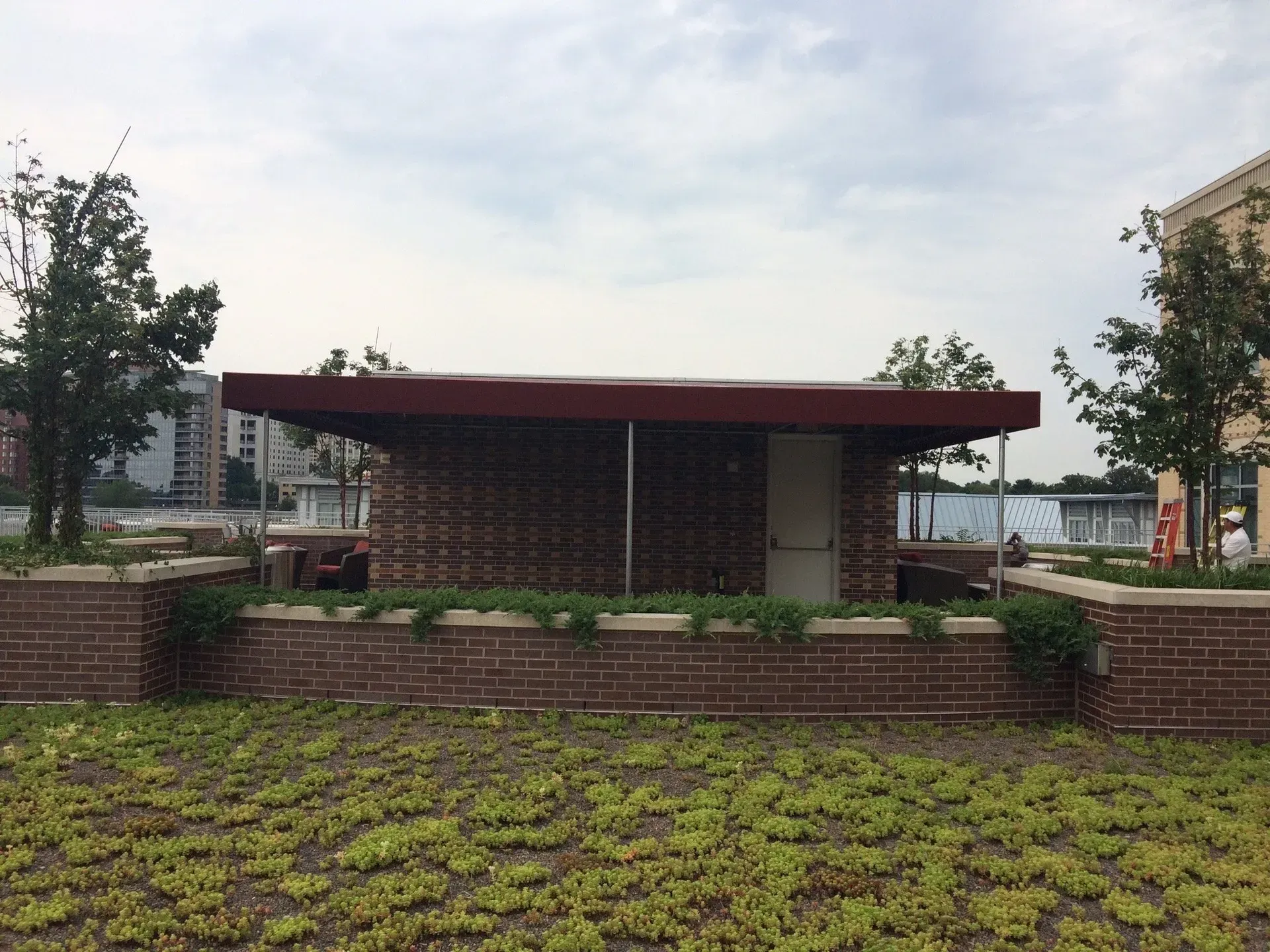 Small brick pavilion with a red roof in a landscaped park, surrounded by grass and trees.