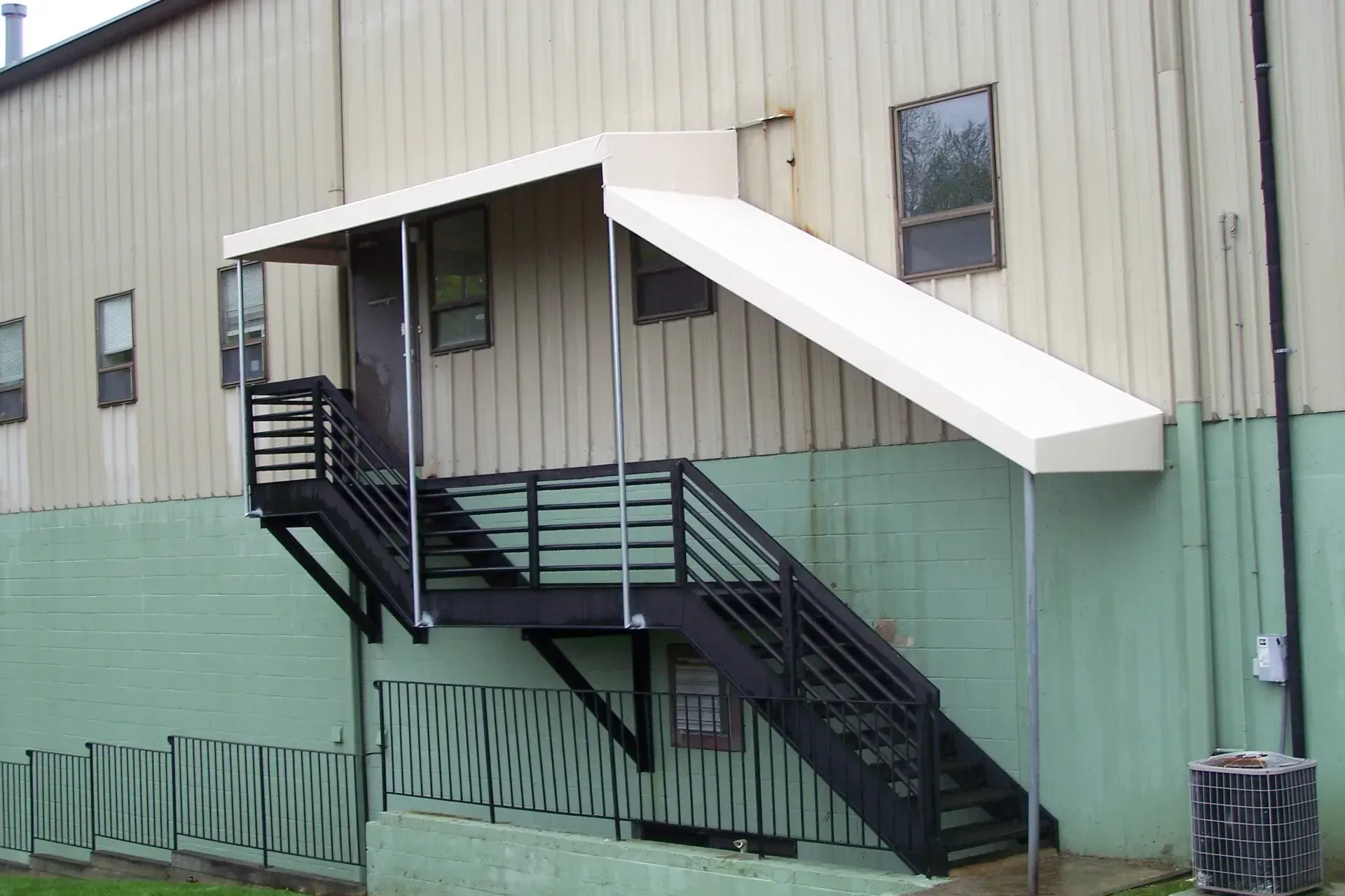Exterior metal stairway and covered entrance on a beige industrial building with green lower siding.