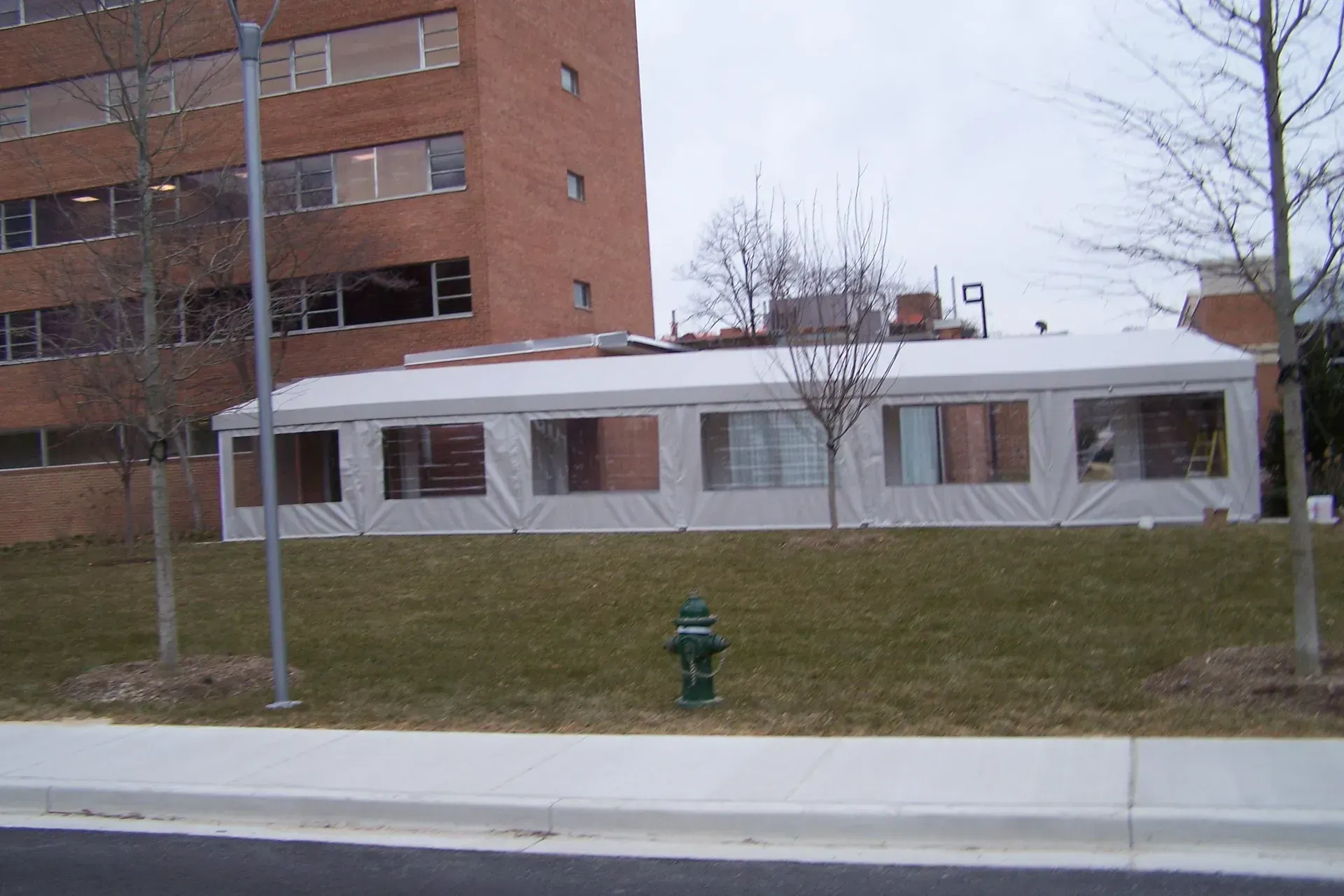 White modular building beside a brick apartment block, with a sidewalk, grassy slope, and fire hydrant.