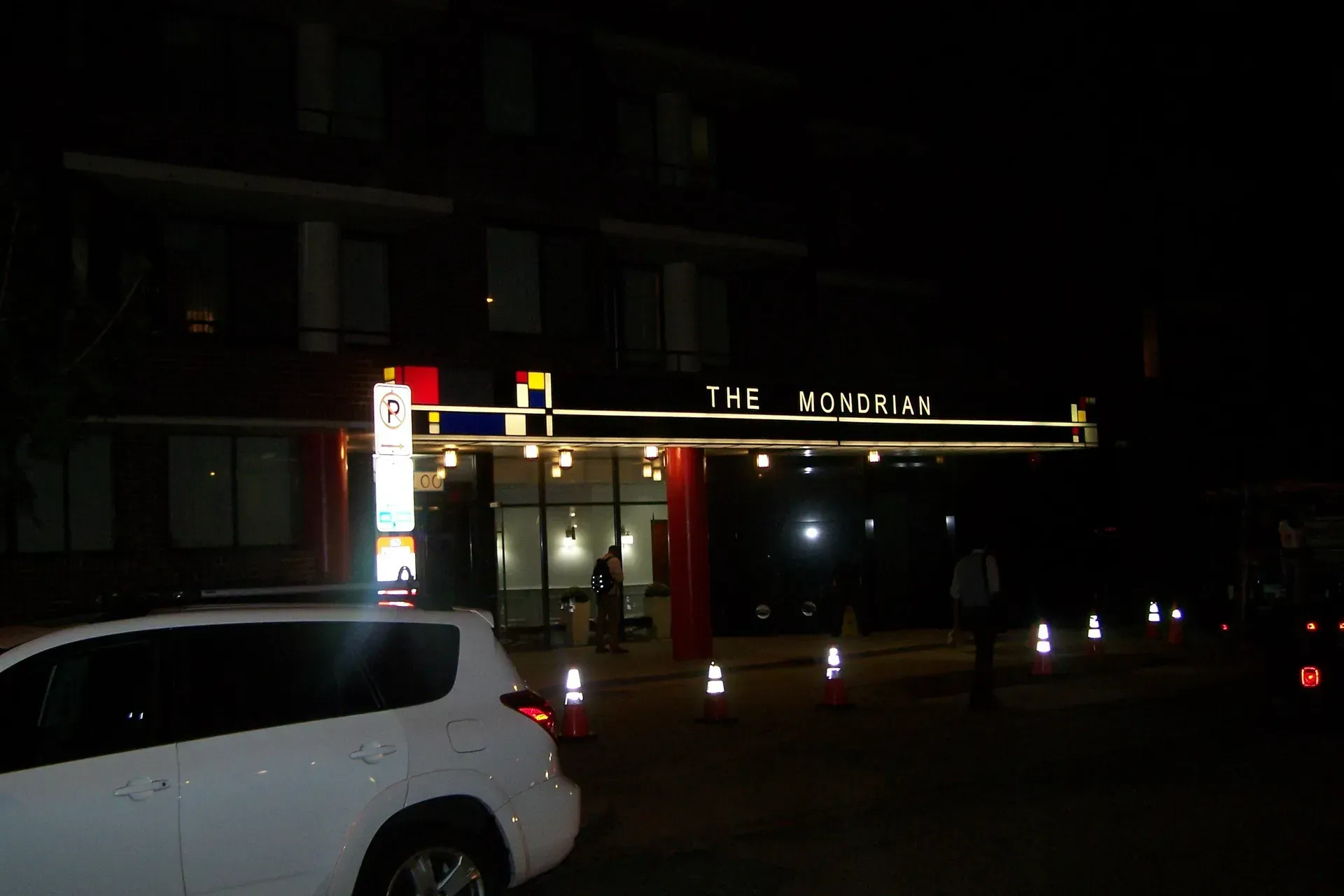 Nighttime street view of a lit restaurant entrance with parked cars and traffic cones