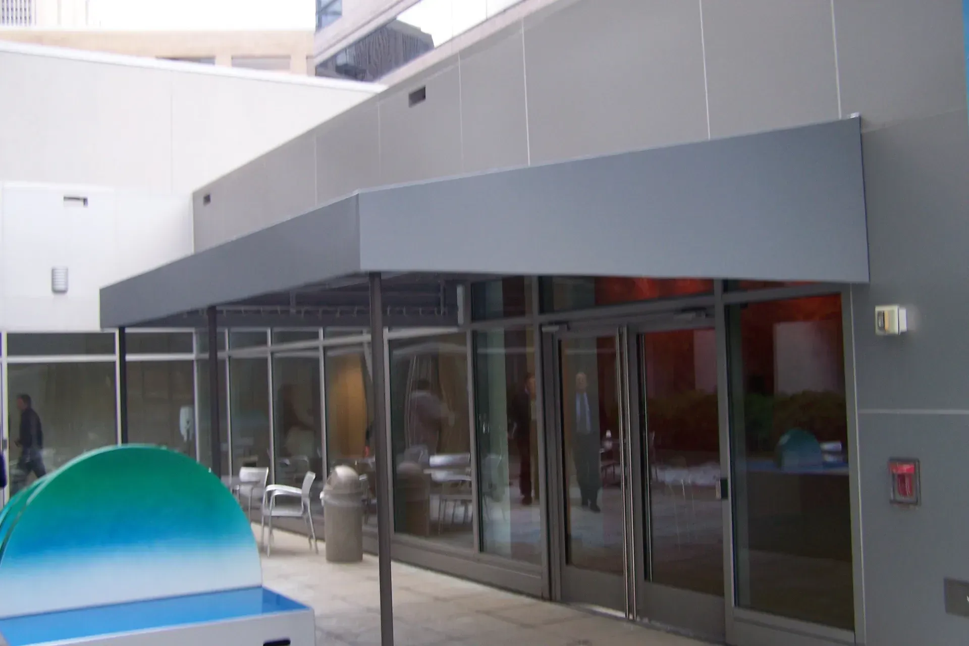 Modern building entrance with gray awning and glass doors, beside a blue curved bench or sculpture
