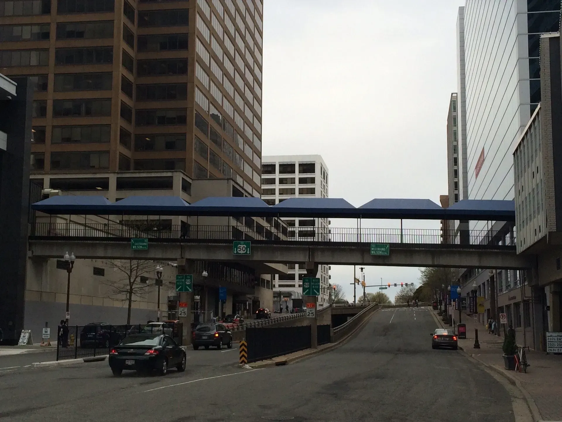 Downtown street with a pedestrian skybridge between tall buildings and light traffic below