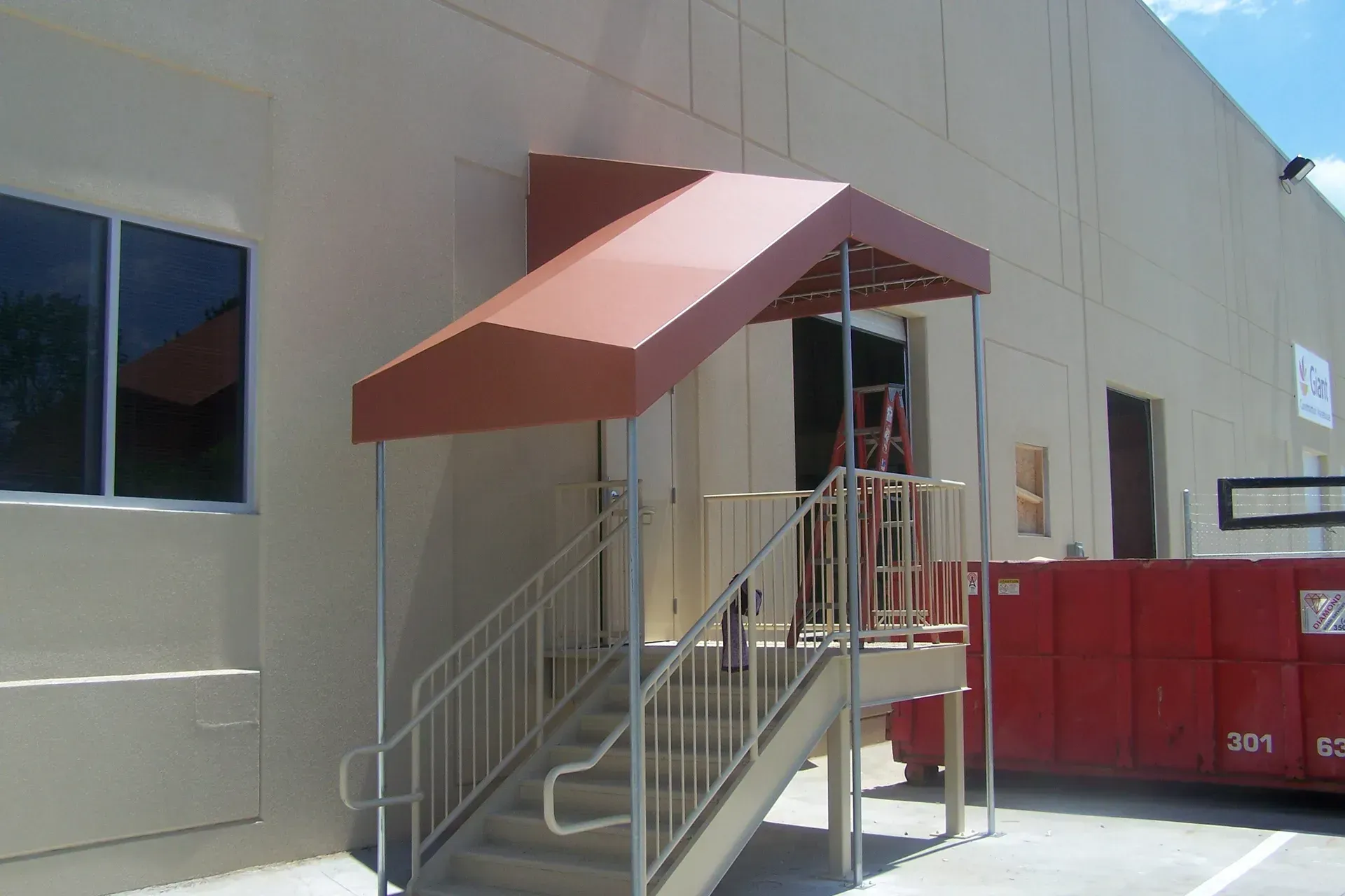 Building entrance with red awning, stairs, and metal handrails beside a red dumpster.