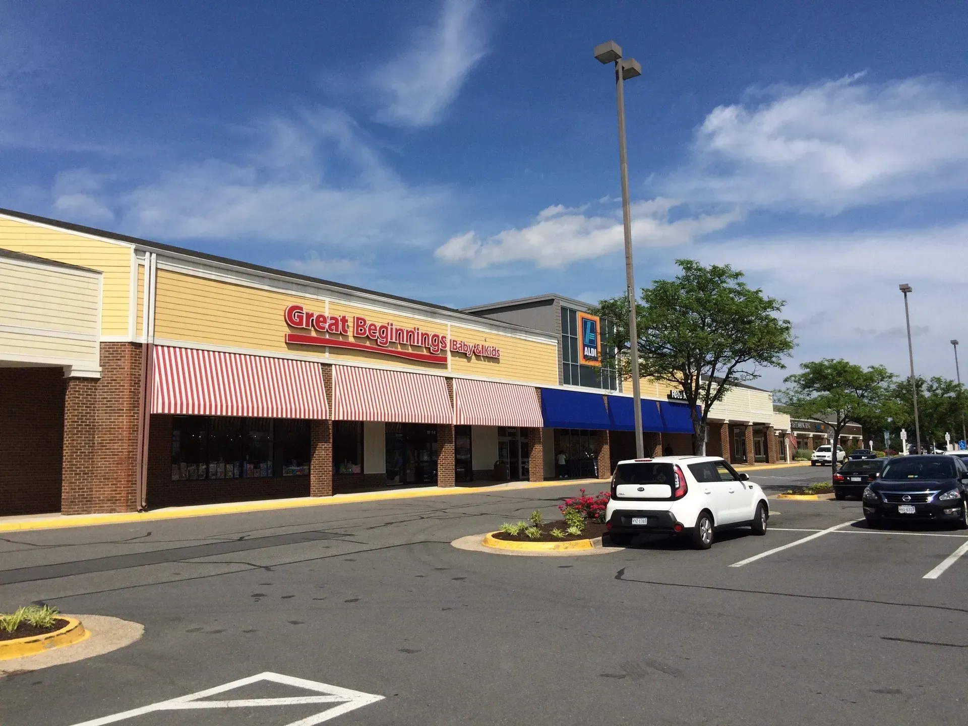 Strip mall storefronts with red and blue awnings beside a parking lot on a sunny day
