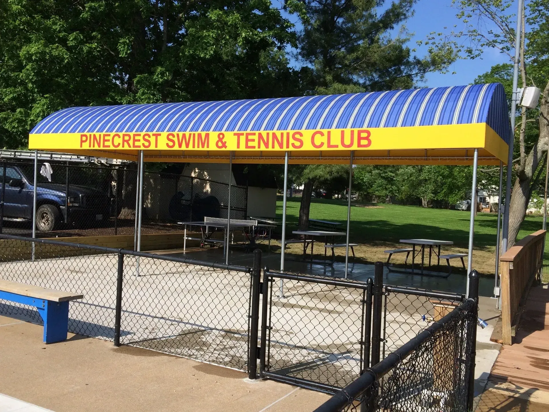 Pinecrest Swim & Tennis Club pavilion with blue-striped roof and yellow sign over fenced seating area