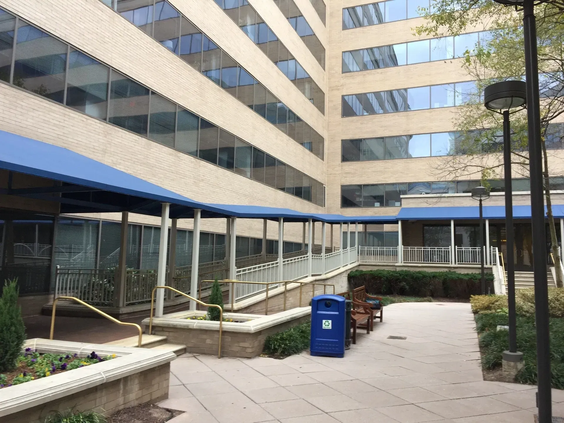 Courtyard between office buildings with glass storefronts, benches, and a blue recycling bin.