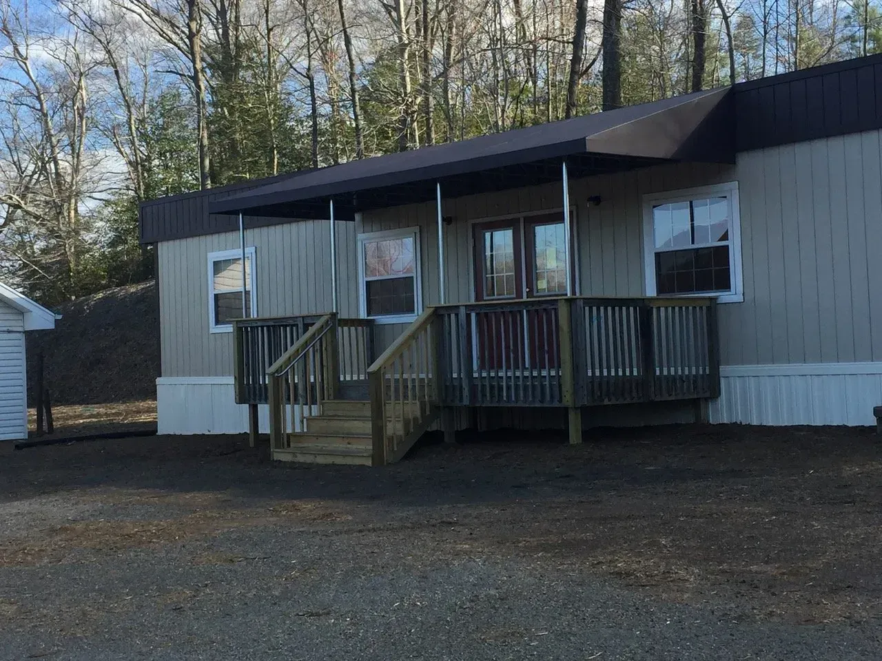 Single-story mobile home with a small front porch and steps on a gravel lot, surrounded by trees