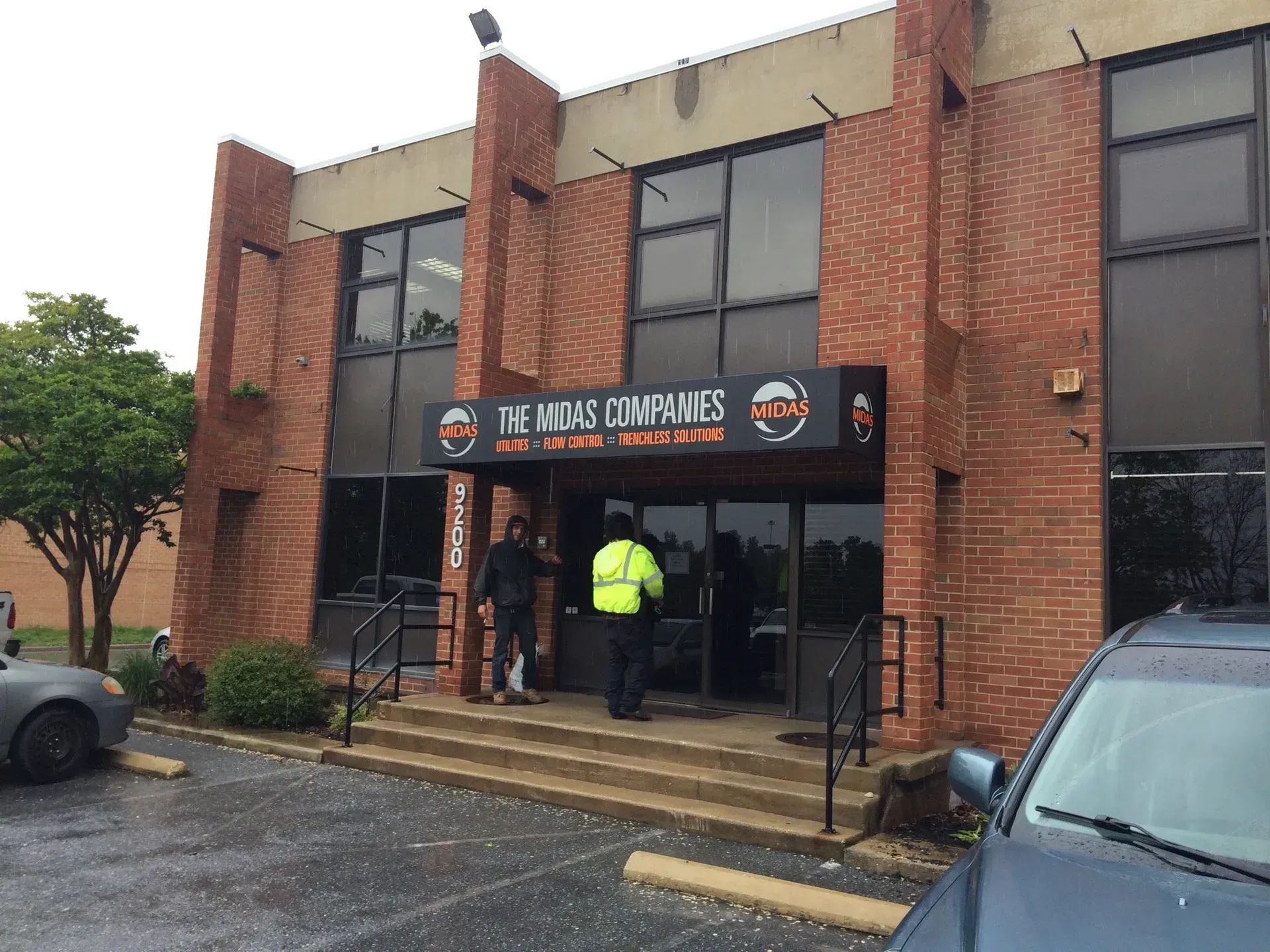Entrance of a brick apartment building with two people near the doorway and parked cars in front