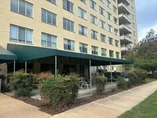 Apartment building with green awnings, patio seating, and shrubs along a sidewalk