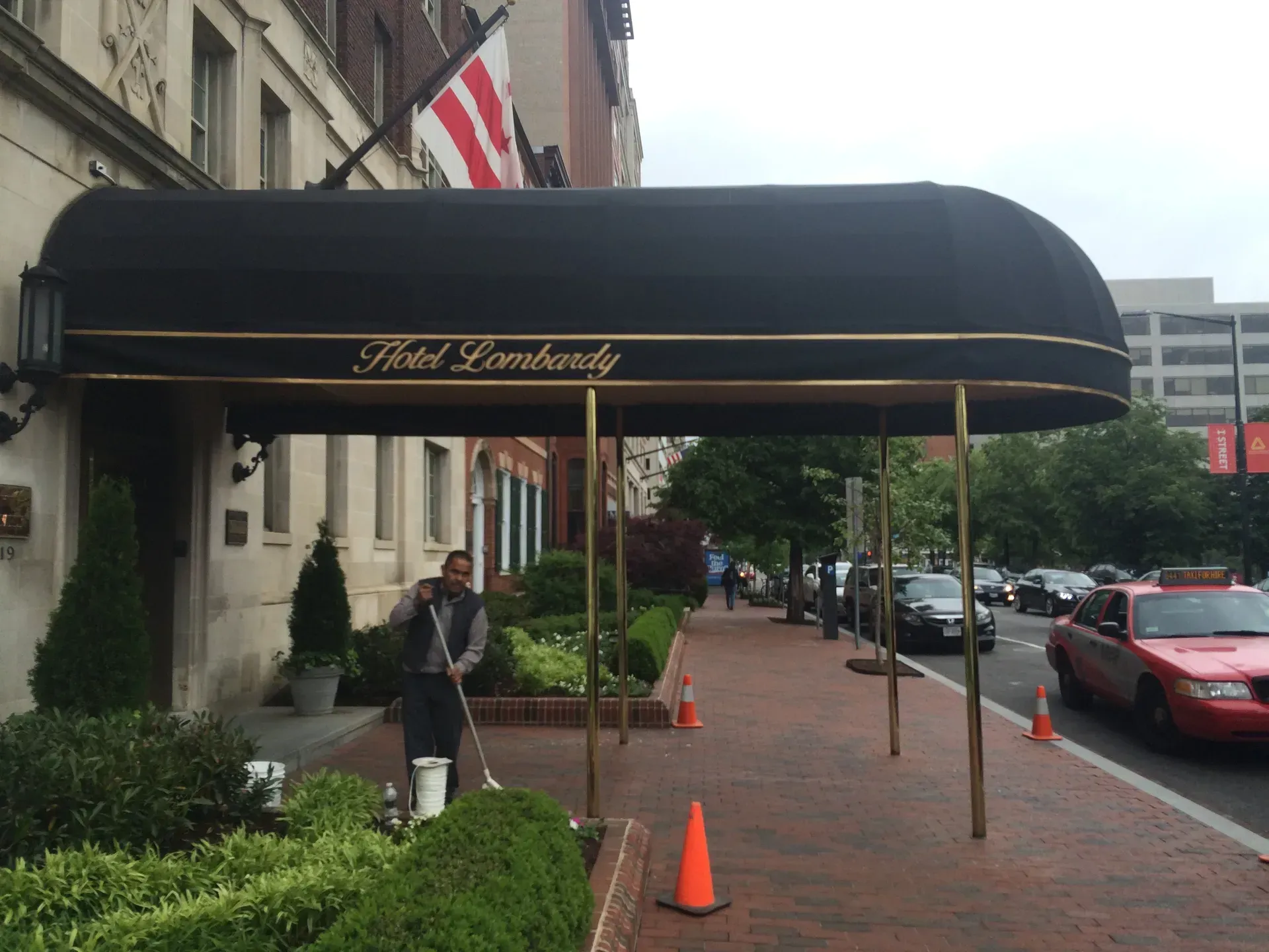 Hotel entrance with black awning, orange cones, and a person standing on a rainy sidewalk