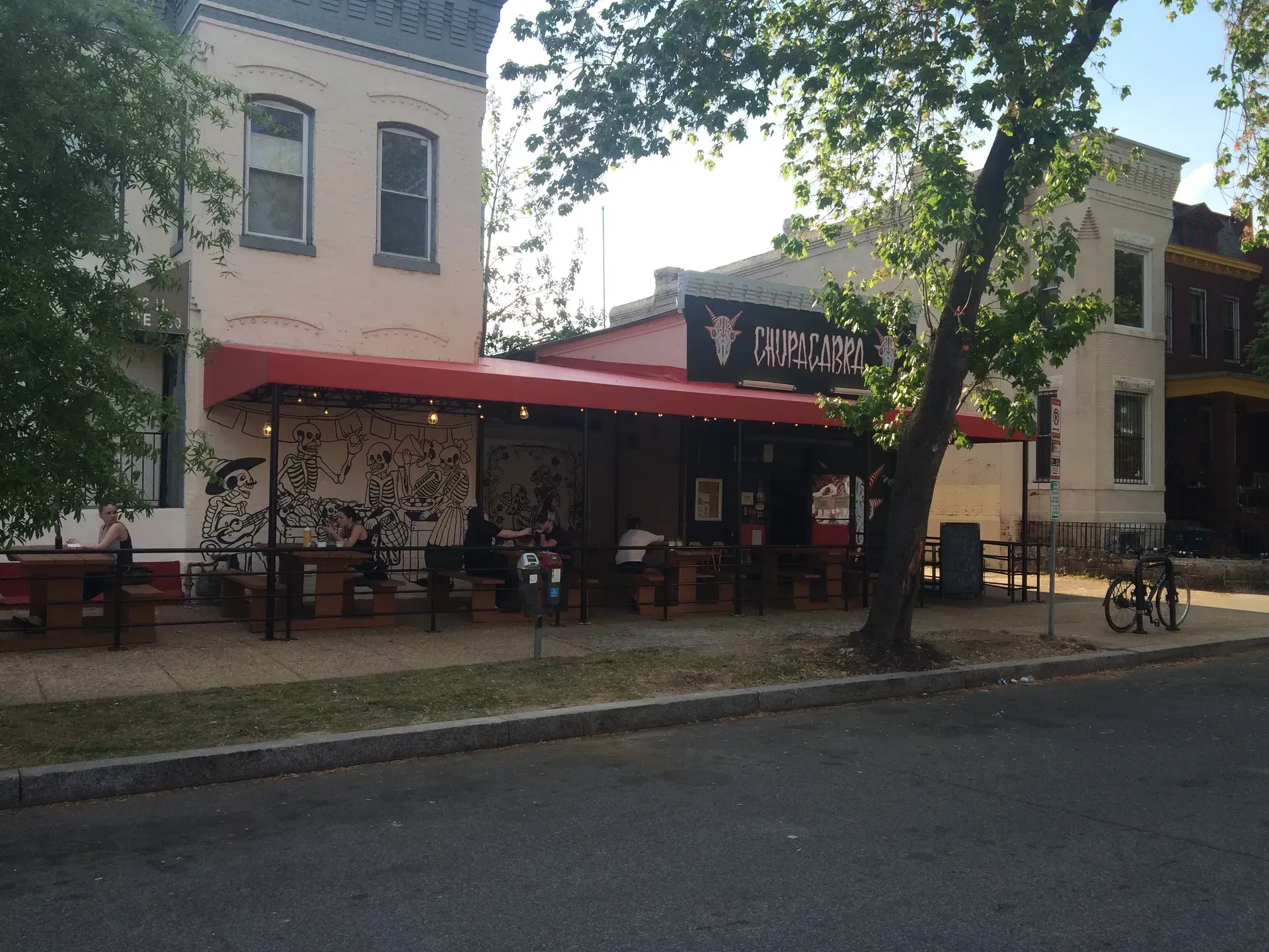 Street view of a café with a red awning and outdoor seating beside a tree-lined sidewalk
