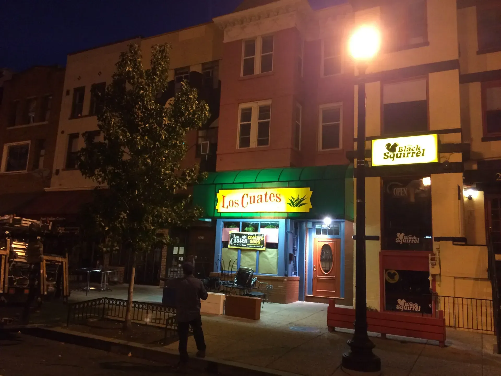 Night street view of a lit storefront with a person walking past under a streetlamp.