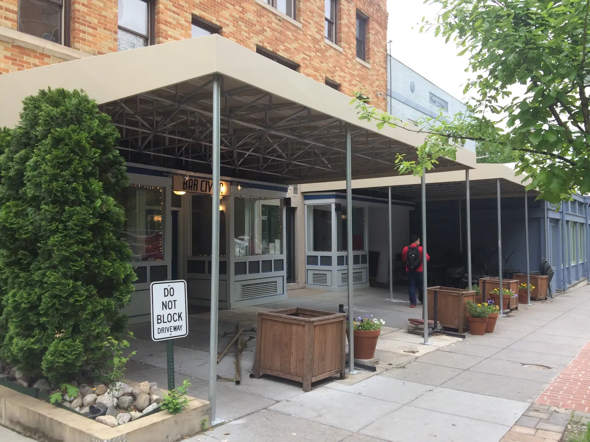 Covered outdoor storefront with potted plants, sidewalk seating, and a “No Dogs Allowed” sign