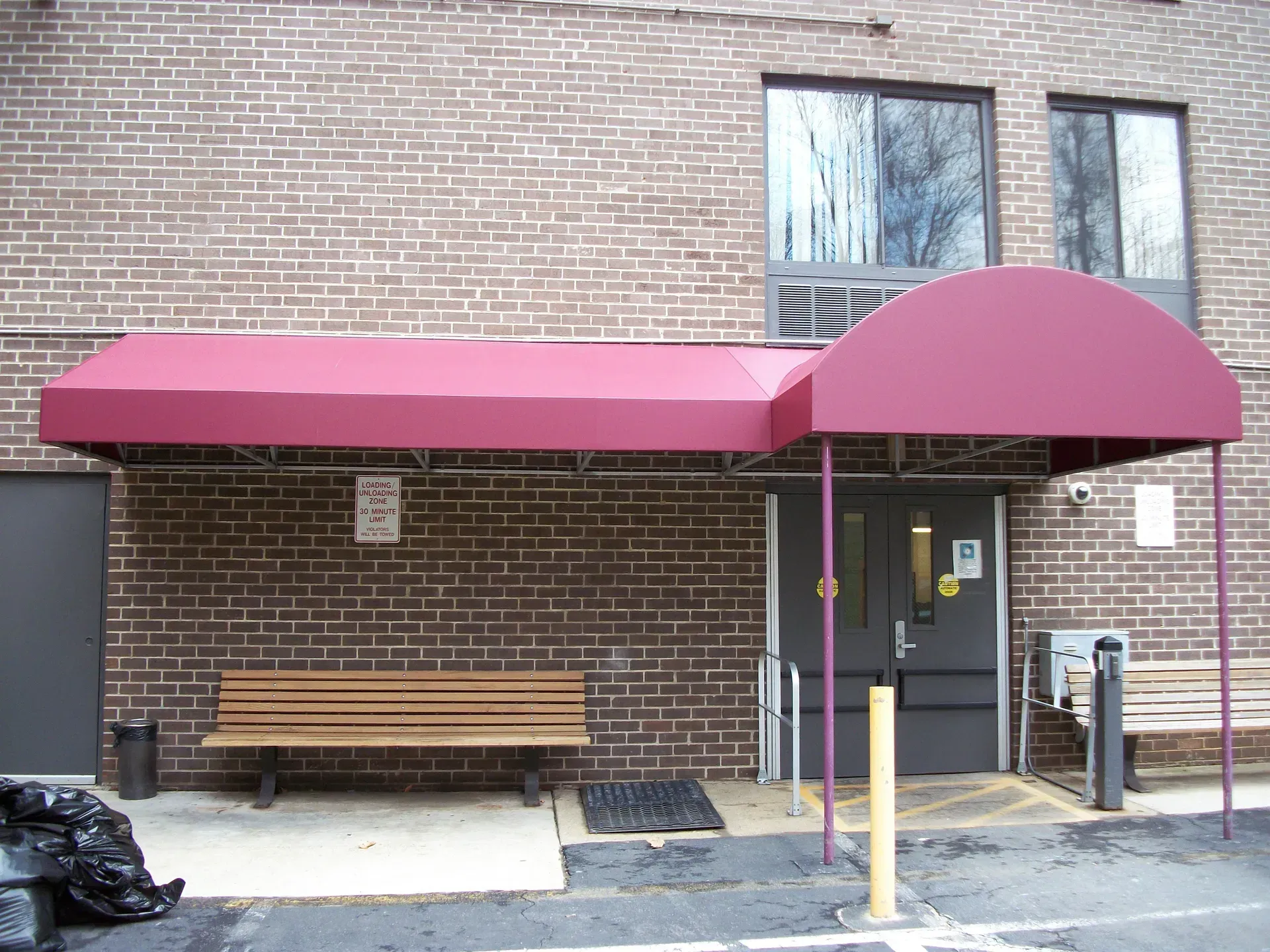 Brick building entrance with maroon awning, bench, and trash bags on the sidewalk