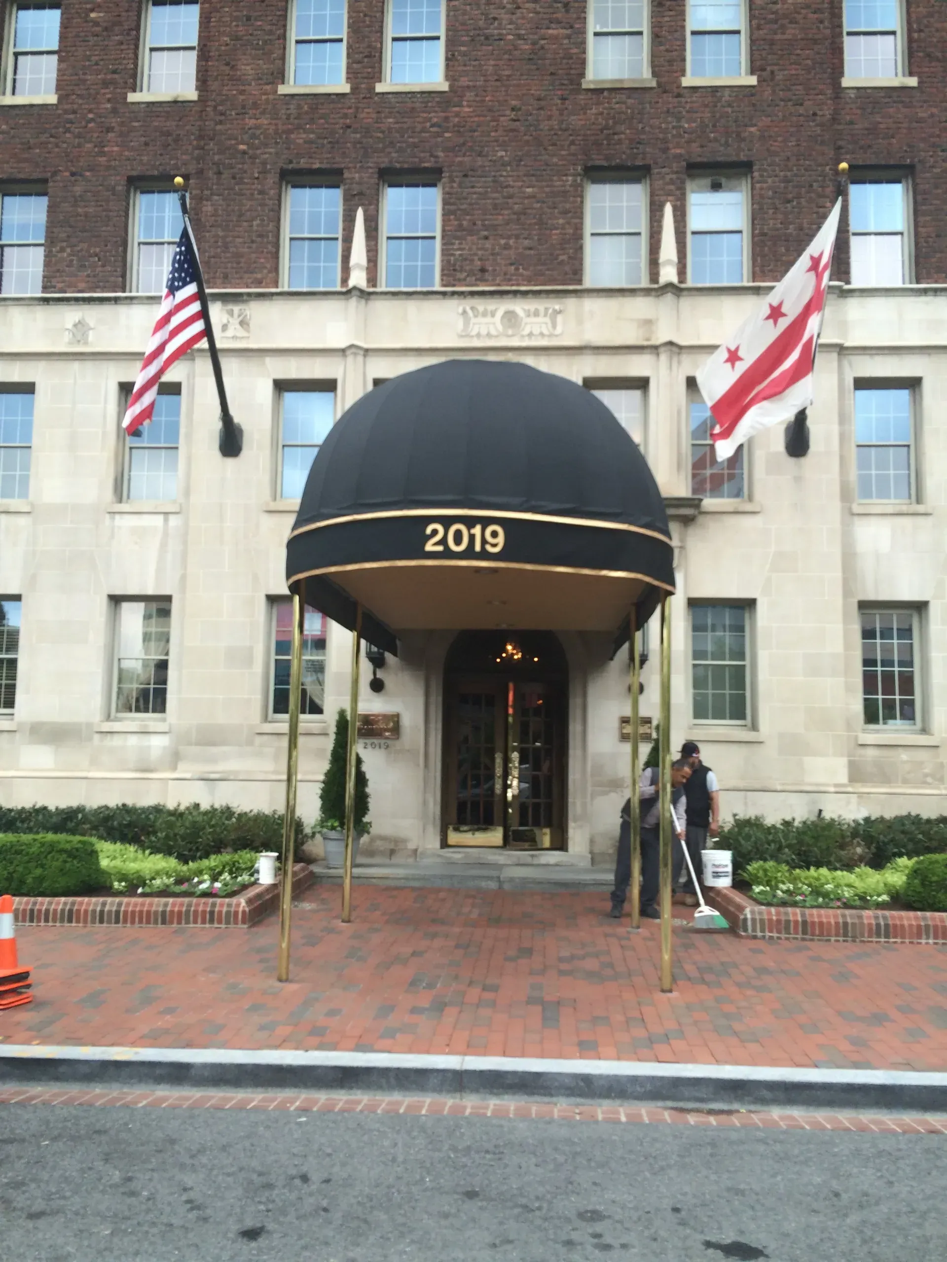 Brick building entrance with black awning marked 2019 and two flags on either side.