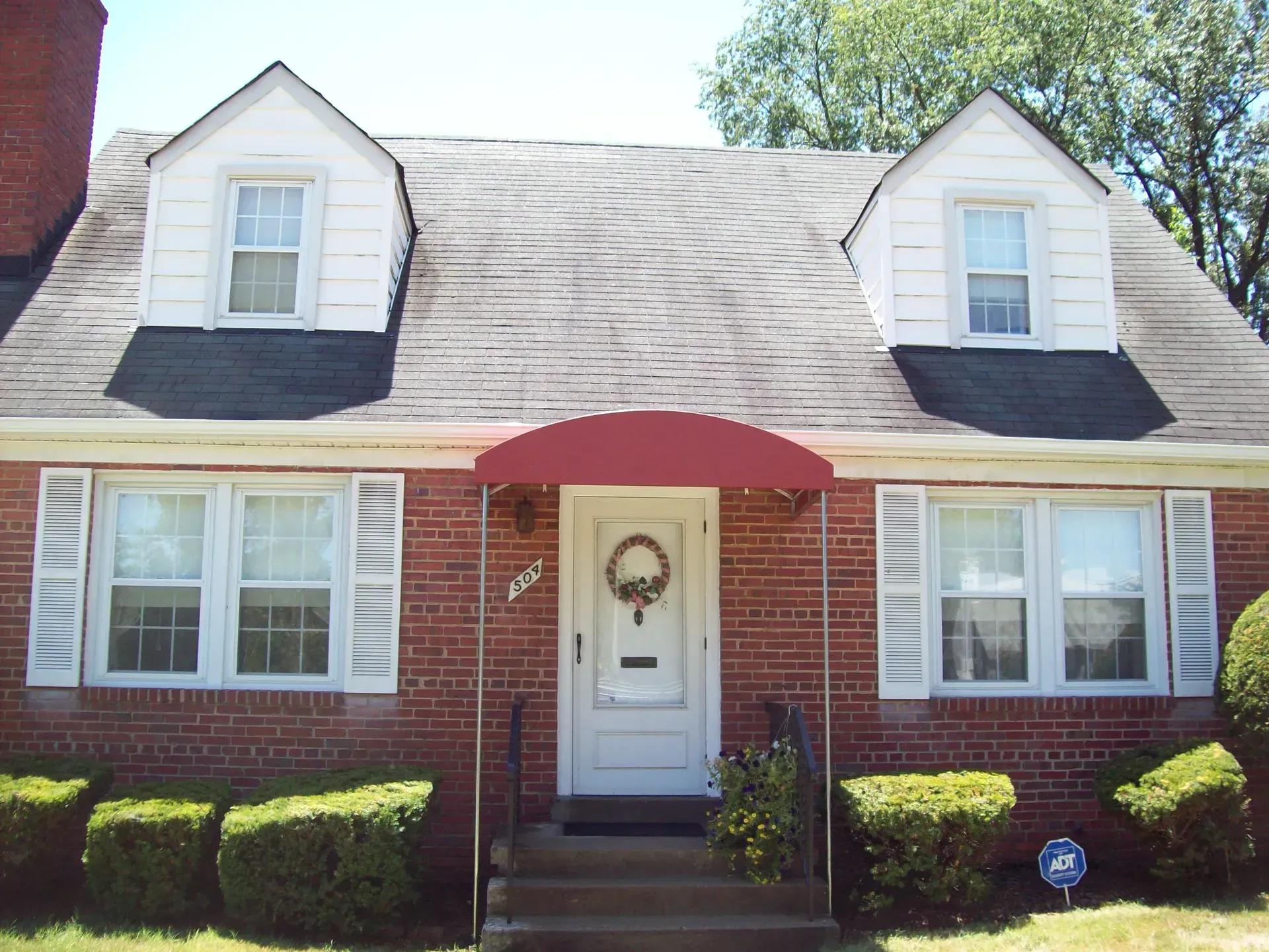 Brick house with white shutters, dormer windows, and a red awning over the front door