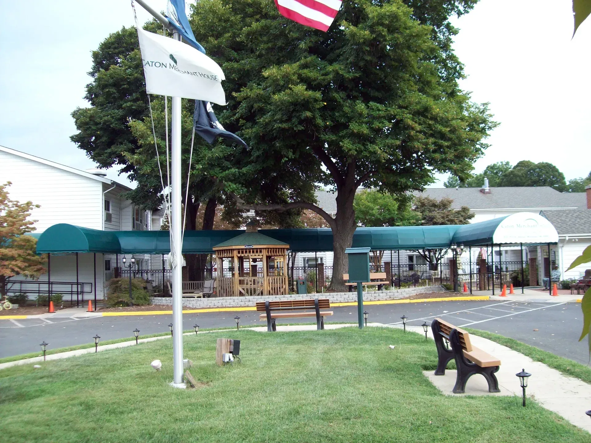 Small park with a flagpole, benches, trees, and a building with a teal awning near a sidewalk.