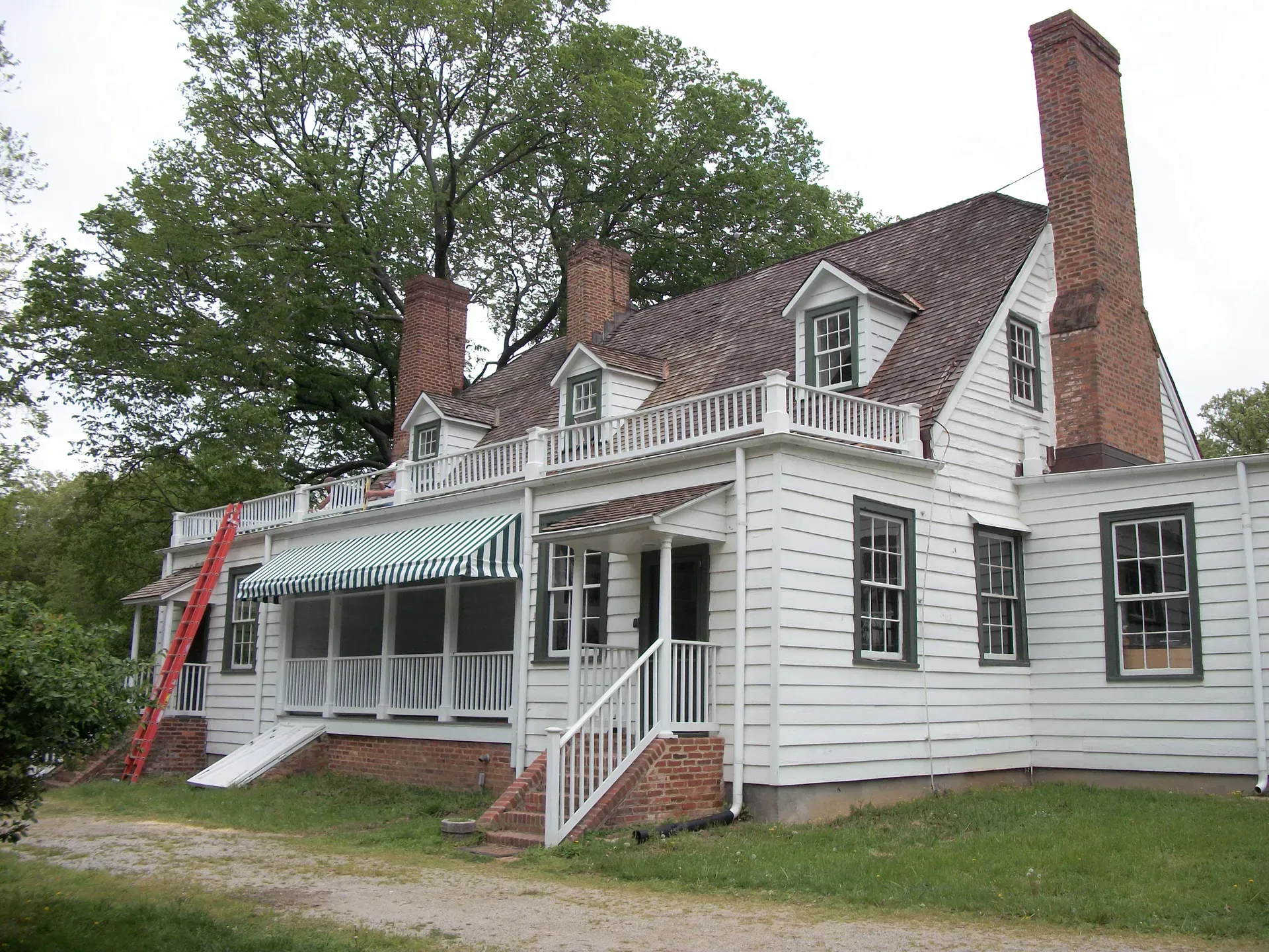 White house with red roof, chimney, and striped awning, set among trees with a ladder on the side