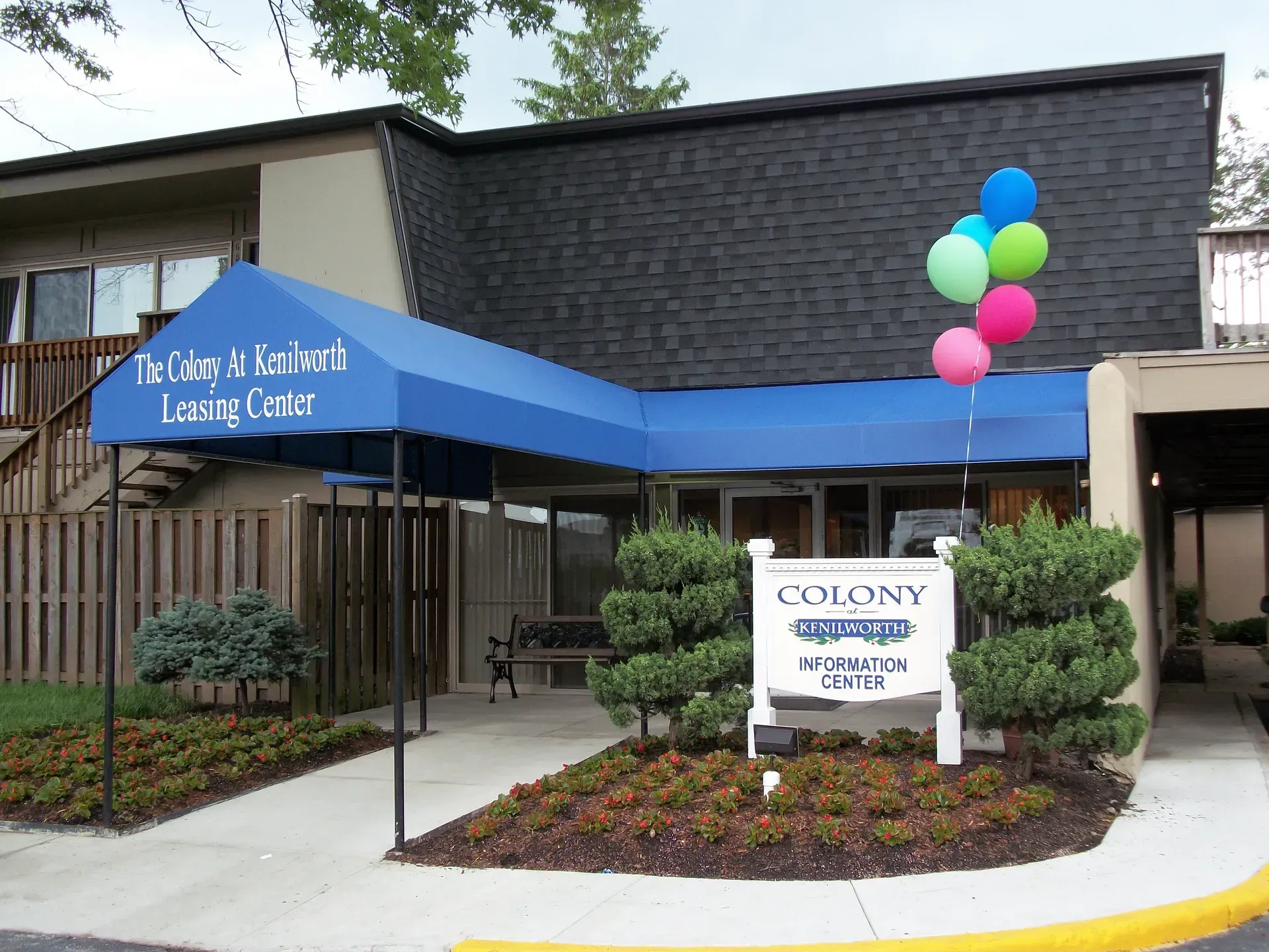 Blue canopy at retirement community entrance with balloons and a sign on a landscaped walkway.