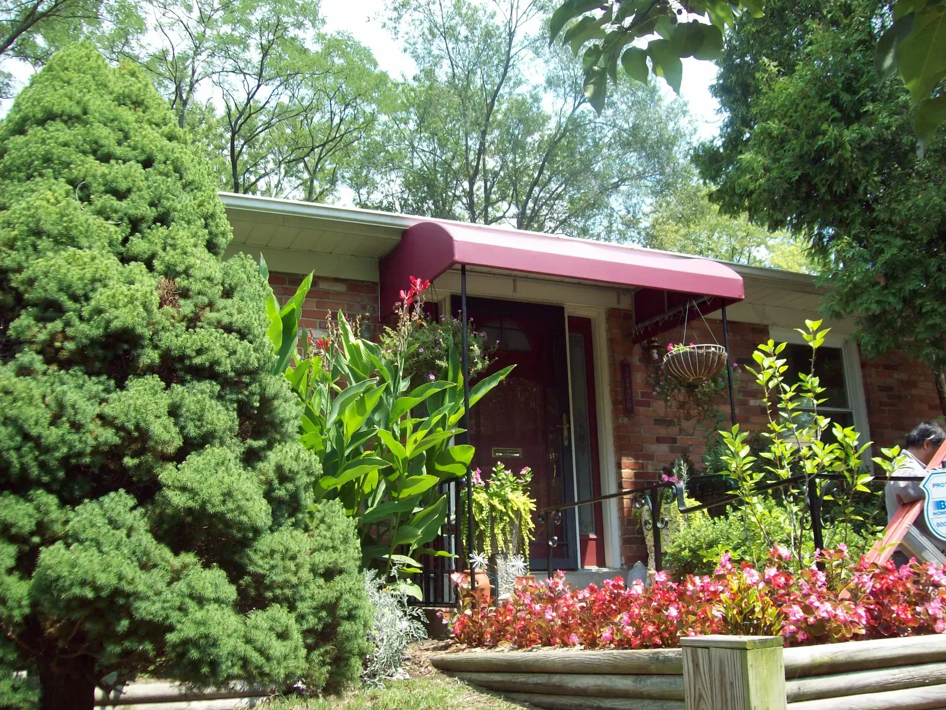 Brick house entrance with red awning, surrounded by lush greenery and colorful flowers.