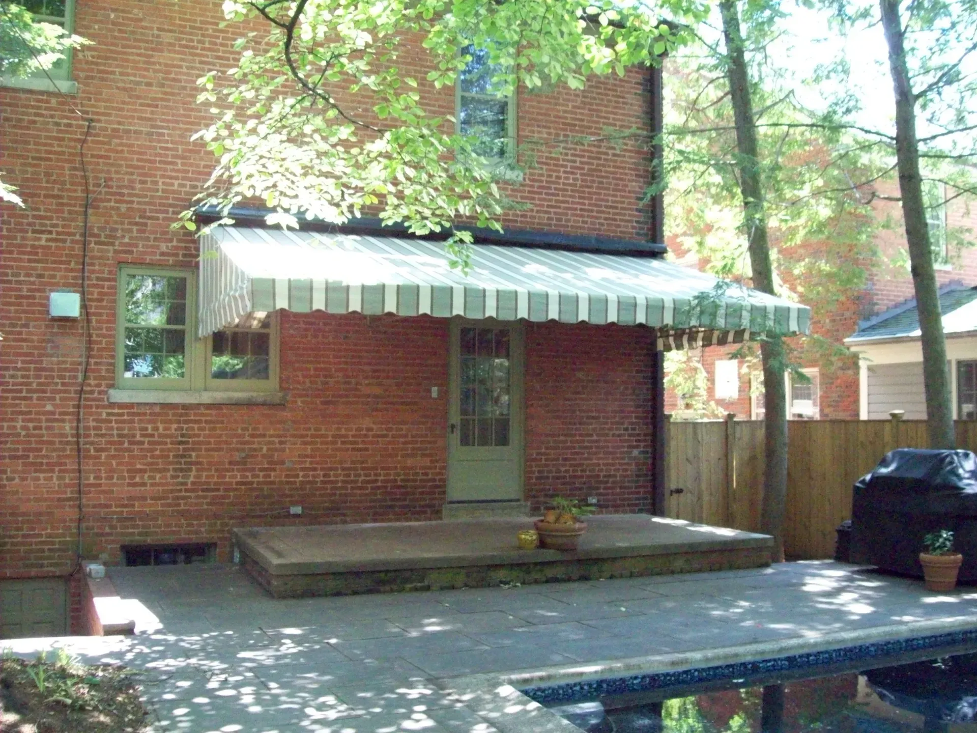 Red brick house with striped awning over a back patio, steps, and a blue tarp-covered pool in the foreground