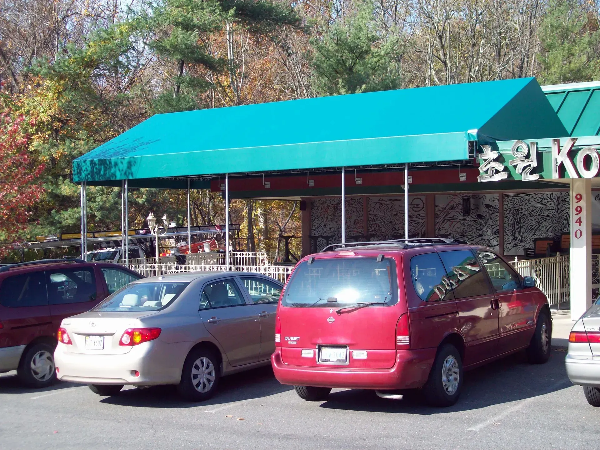 Parking lot with cars in front of a storefront with a teal awning and trees in the background