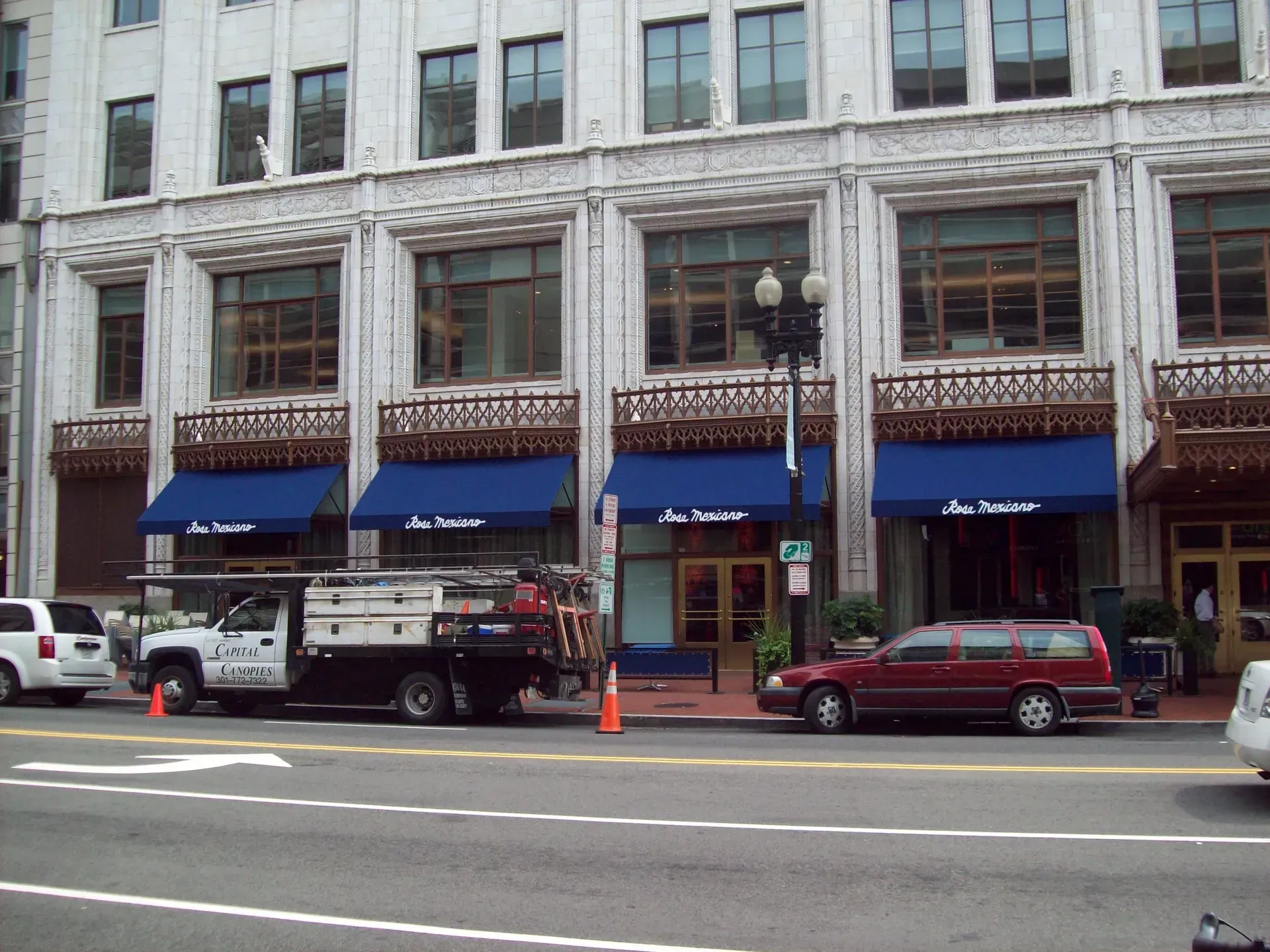 Street view of a stone building with blue awnings, parked trucks and cars along the curb.