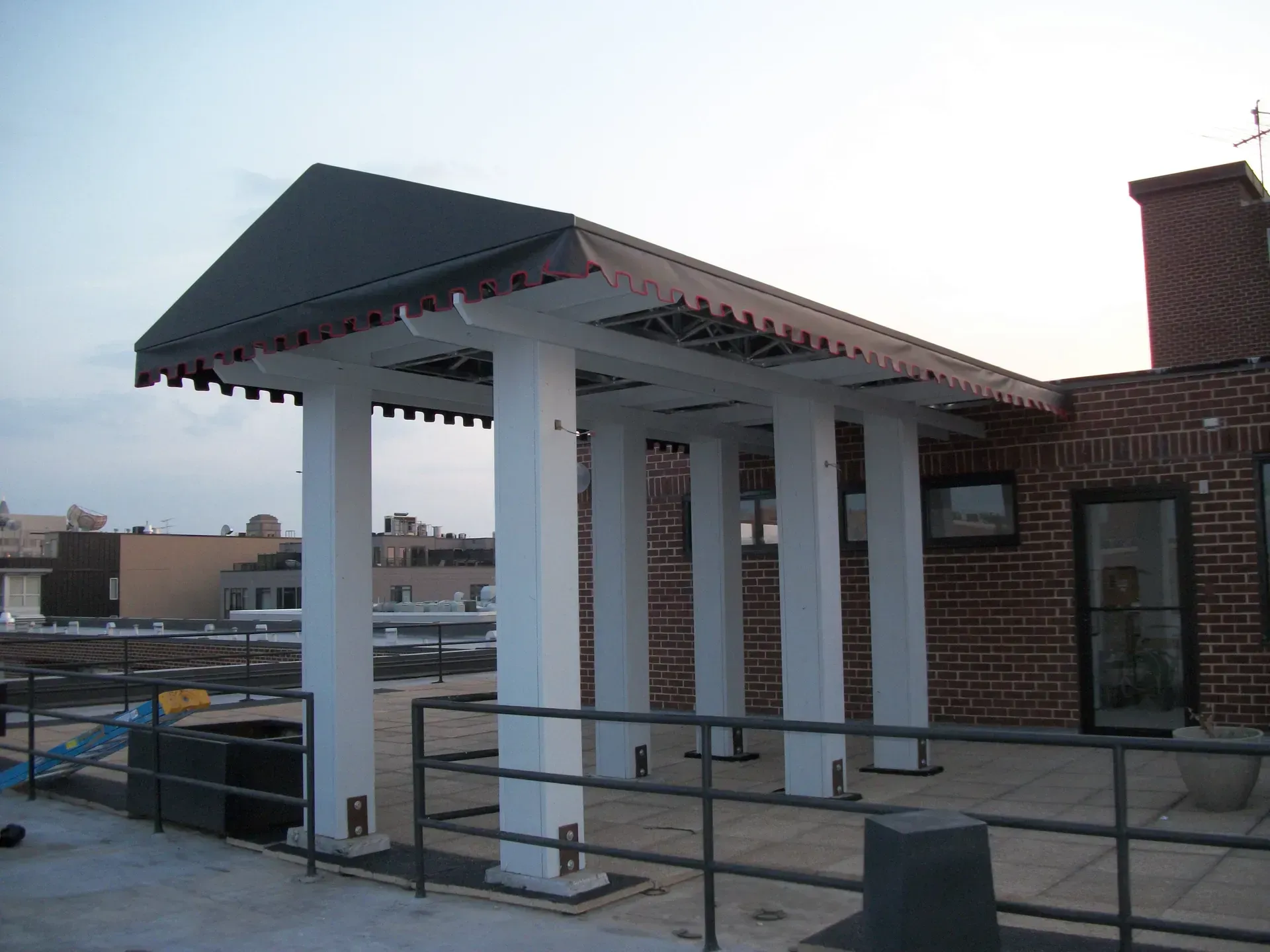 Rooftop walkway with white columns, black railings, and a dark peaked canopy beside a brick building.