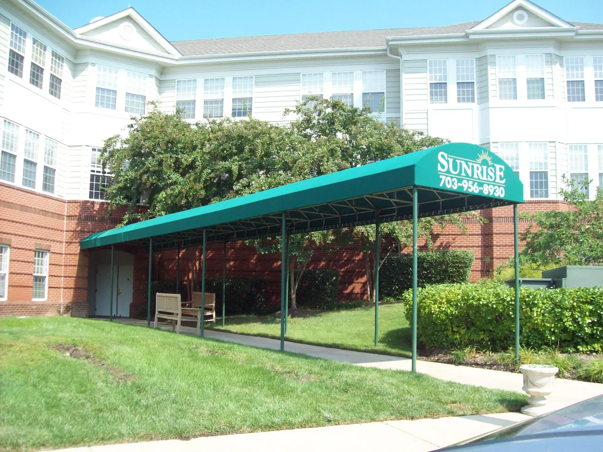 Covered walkway with teal awning beside a brick and white apartment building, with benches and landscaped lawn.