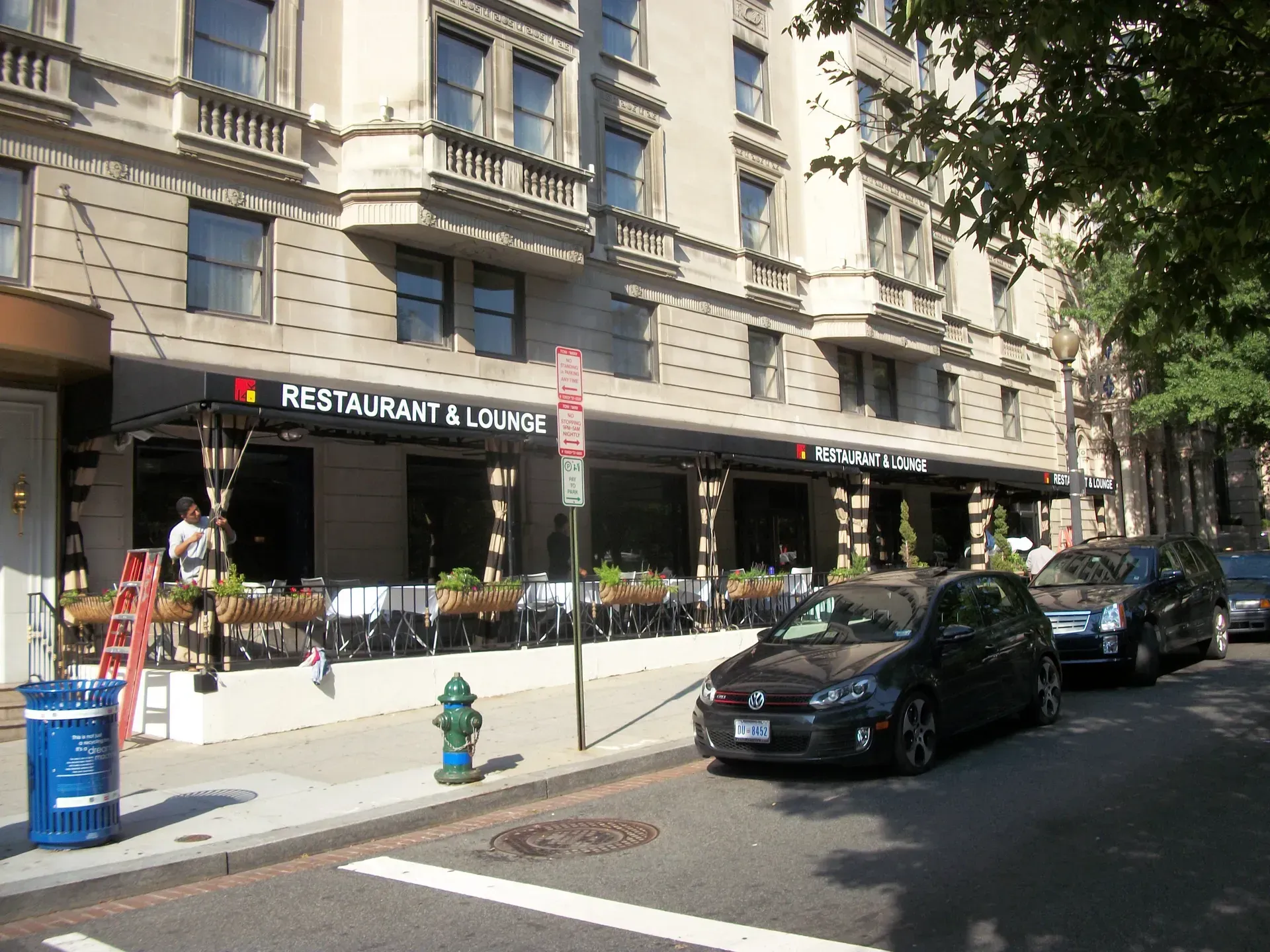 Street-side restaurant with outdoor seating and parked cars along a city block.