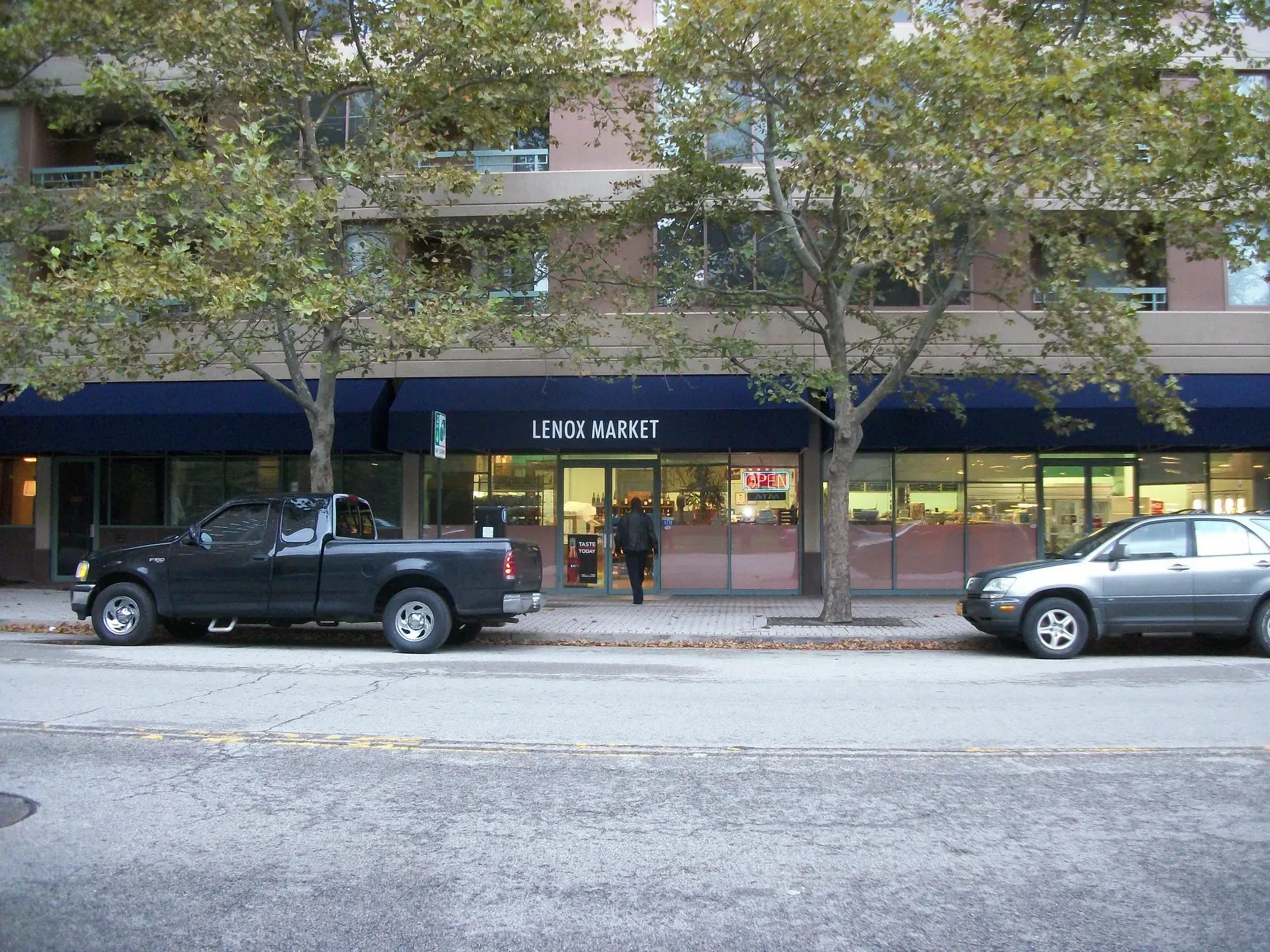 Street view of a storefront with blue awnings, parked black pickup and silver car, and trees lining the sidewalk