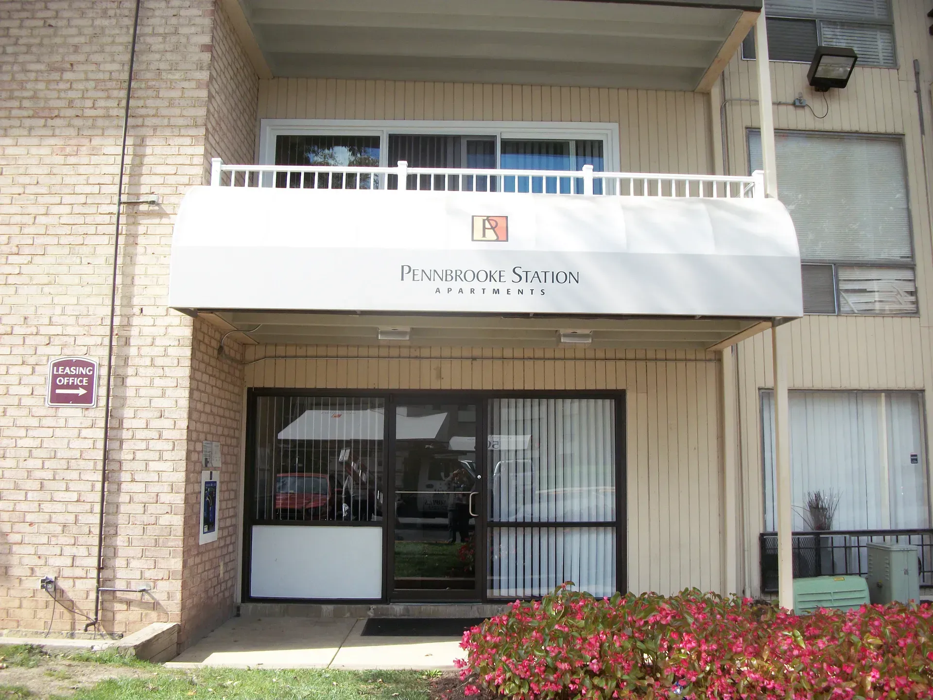 Apartment entrance with white awning, brick facade, glass doors, and flowering bushes.