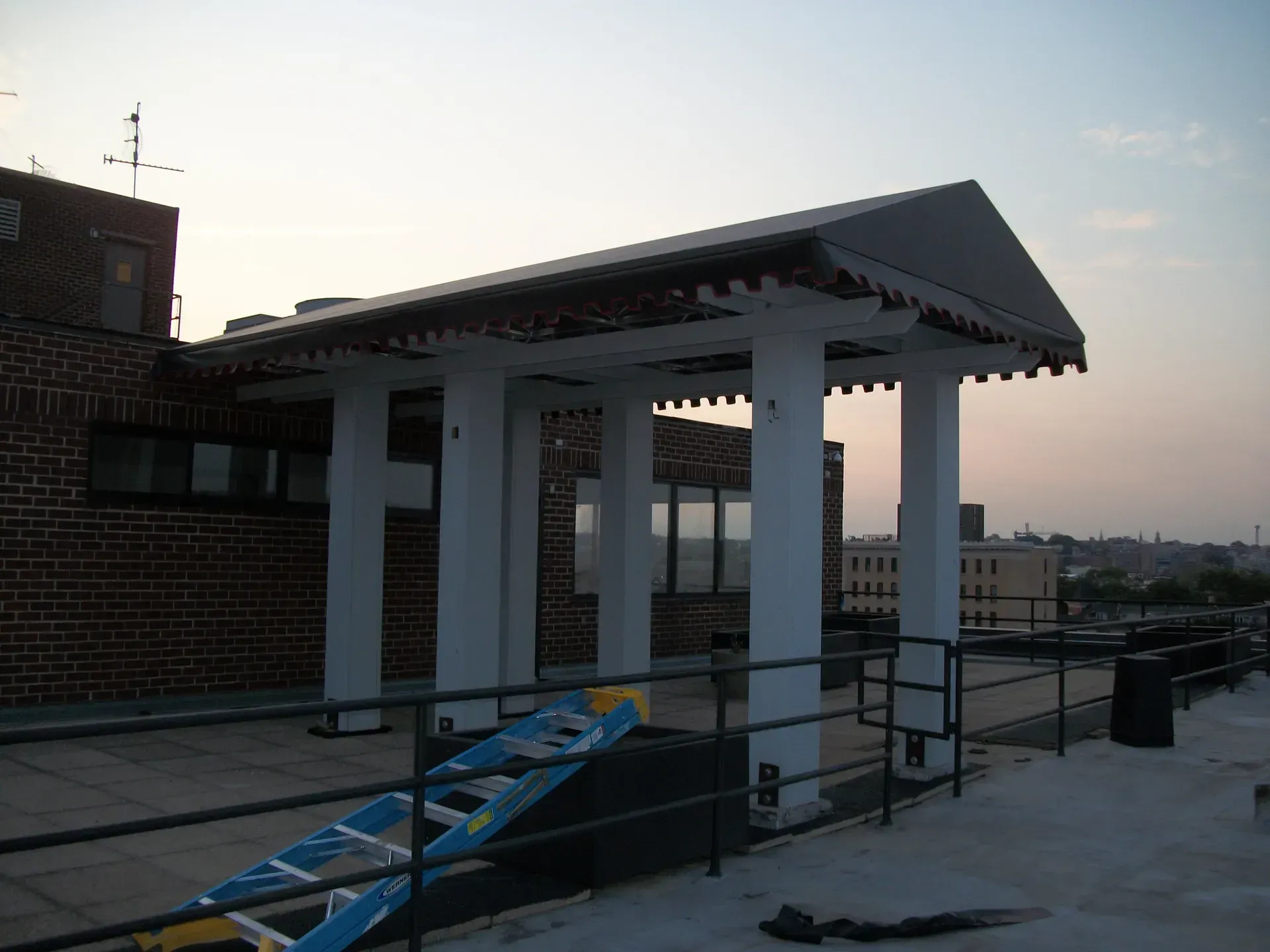 Rooftop walkway with white columns, a sloped roof, and a blue ramp beside a brick building at sunset